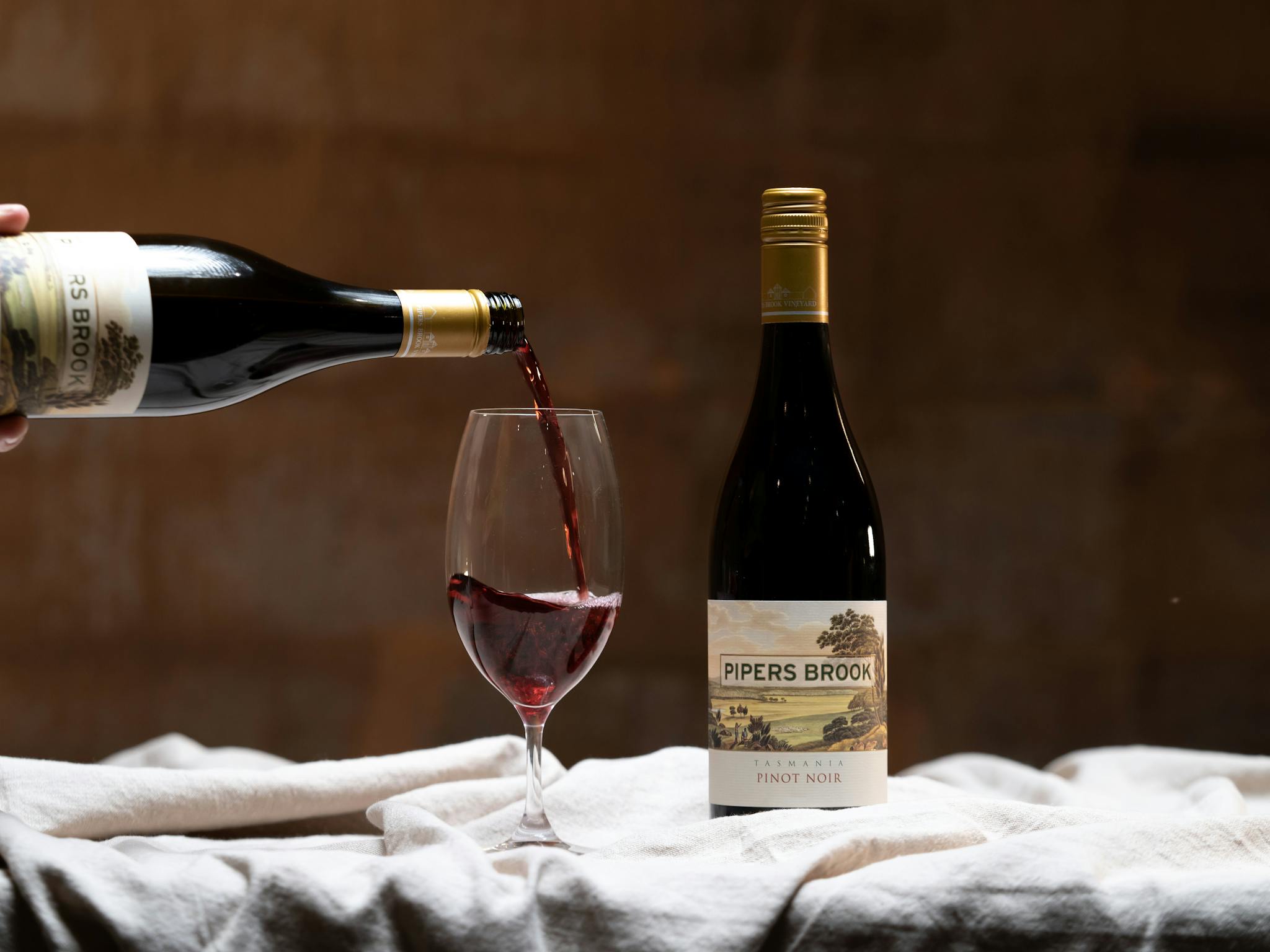 A bottle of Tasmanian Pinot Noir being poured into a glass in a warmly lit cellar.