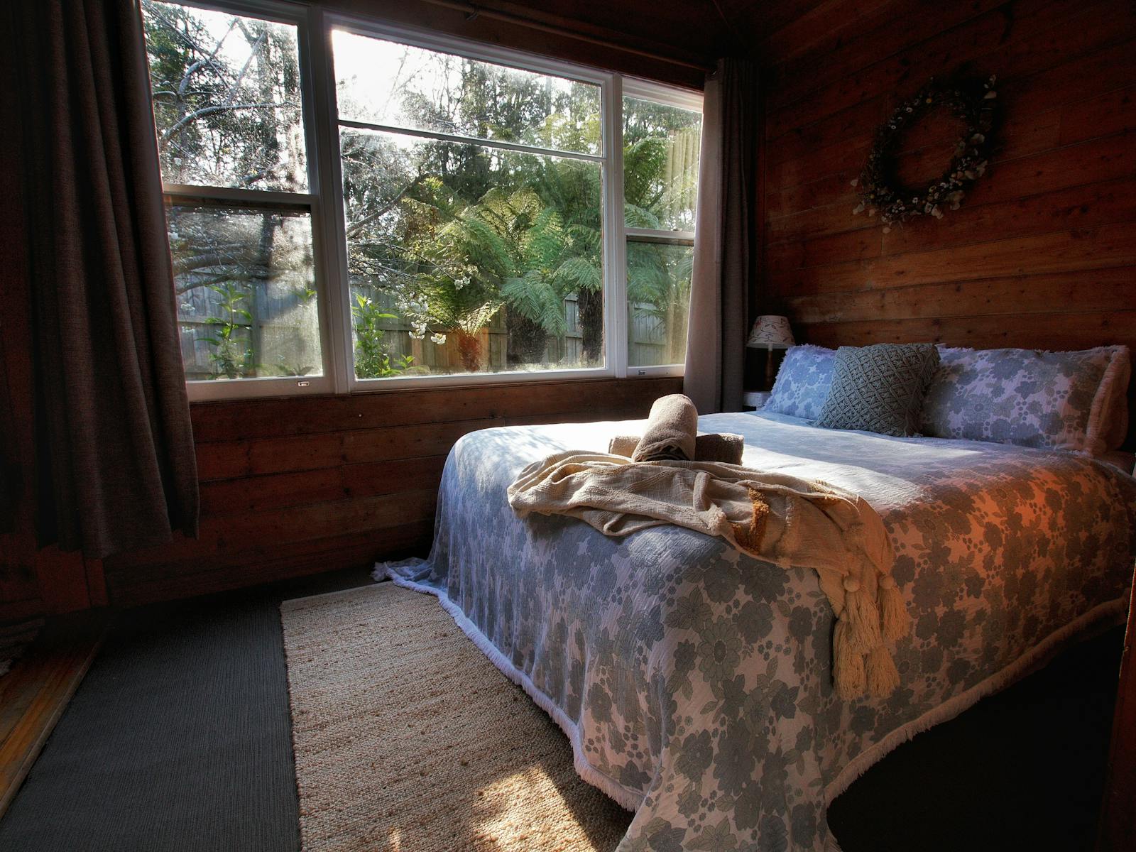 Sun lit main bedroom, the bed draped in a dusty blue cotton spread and fern trees through the window