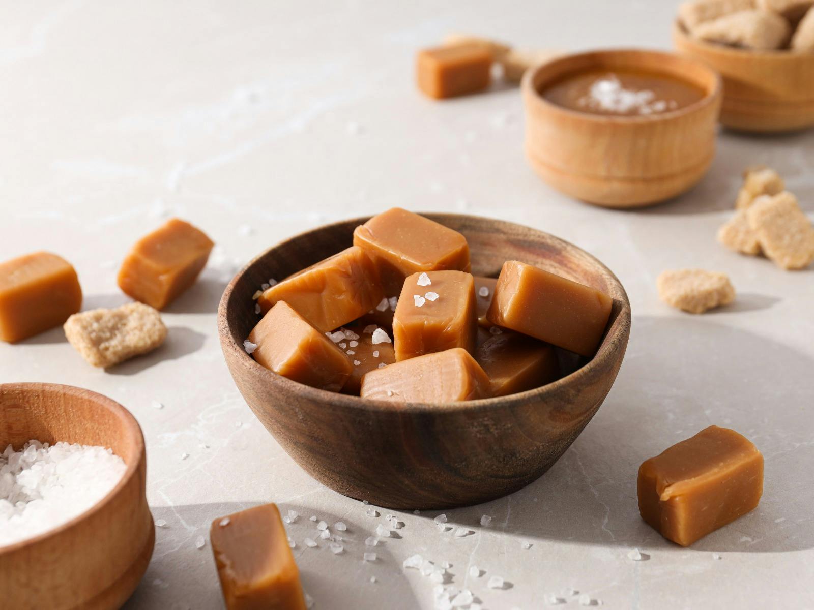 Cubes of salted caramel fudge in a wooden bowl along with sea salt and scattered fudge pieces.