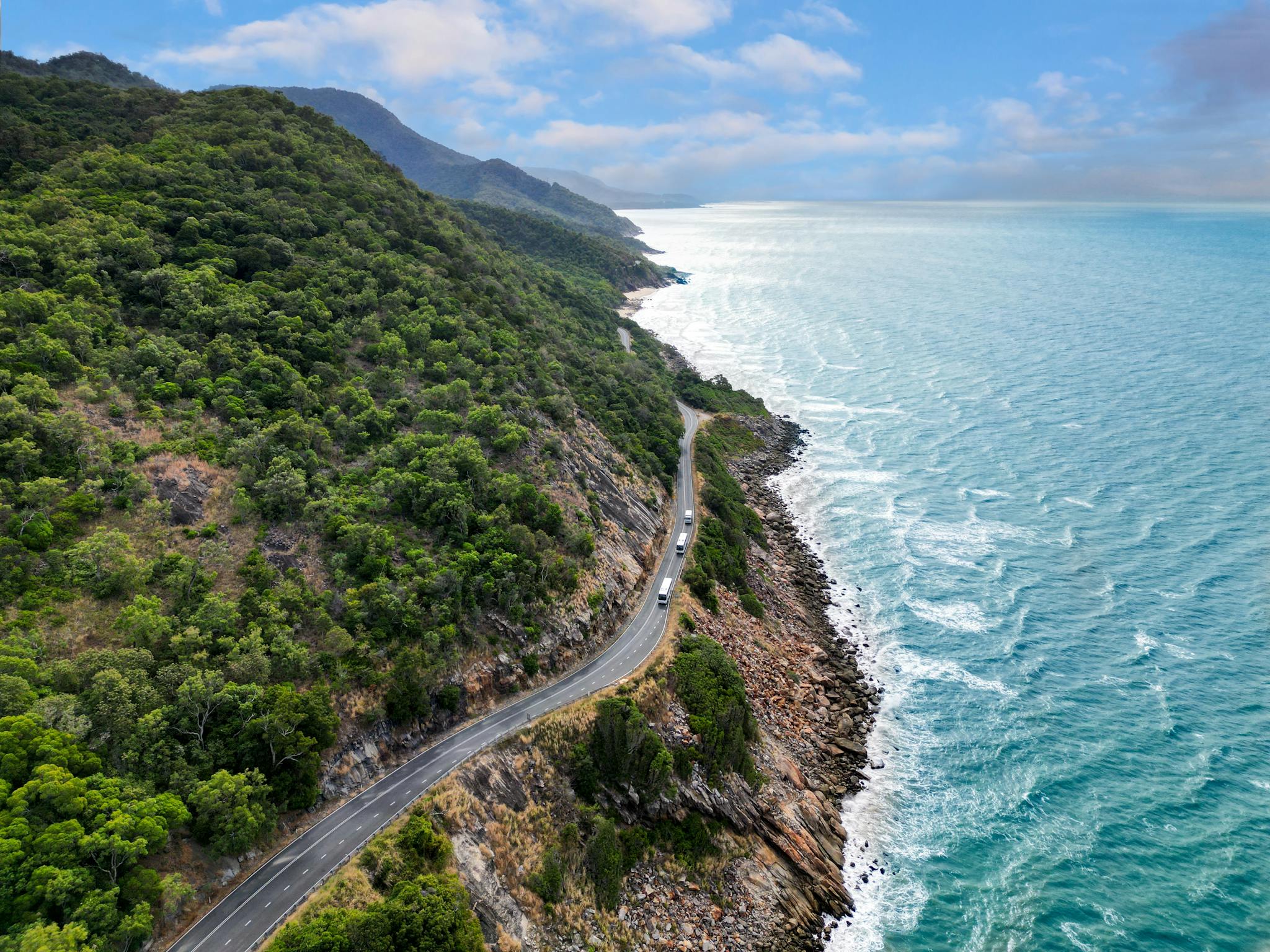 Aerial view of coaches on coastal road with rainforest clad mountains meeting the sea Aerial view of coaches on coastal road with rainforest clad mountains meeting the sea