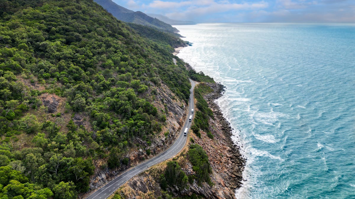 Aerial view of coaches on coastal road with rainforest clad mountains meeting the sea