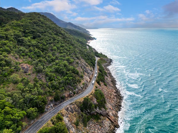 Aerial view of coaches on coastal road with rainforest clad mountains meeting the sea