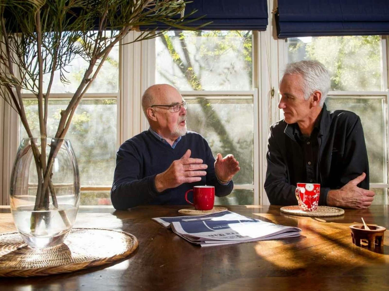 Two men converse at a wooden table near windows, with a vase of branches and mugs.