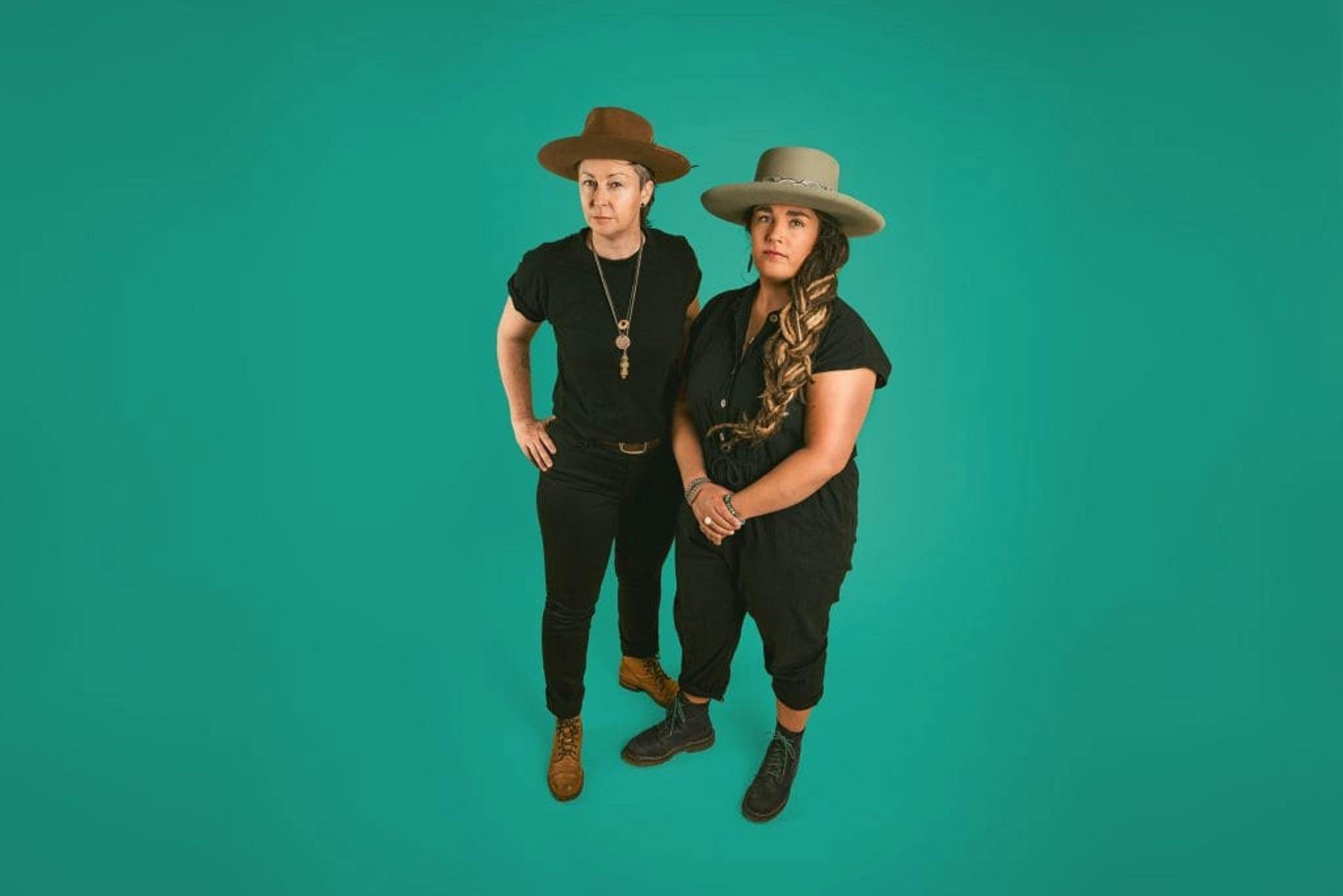 Two musicians standing together wearing all black, and hats. the background is a solid blue-greenish