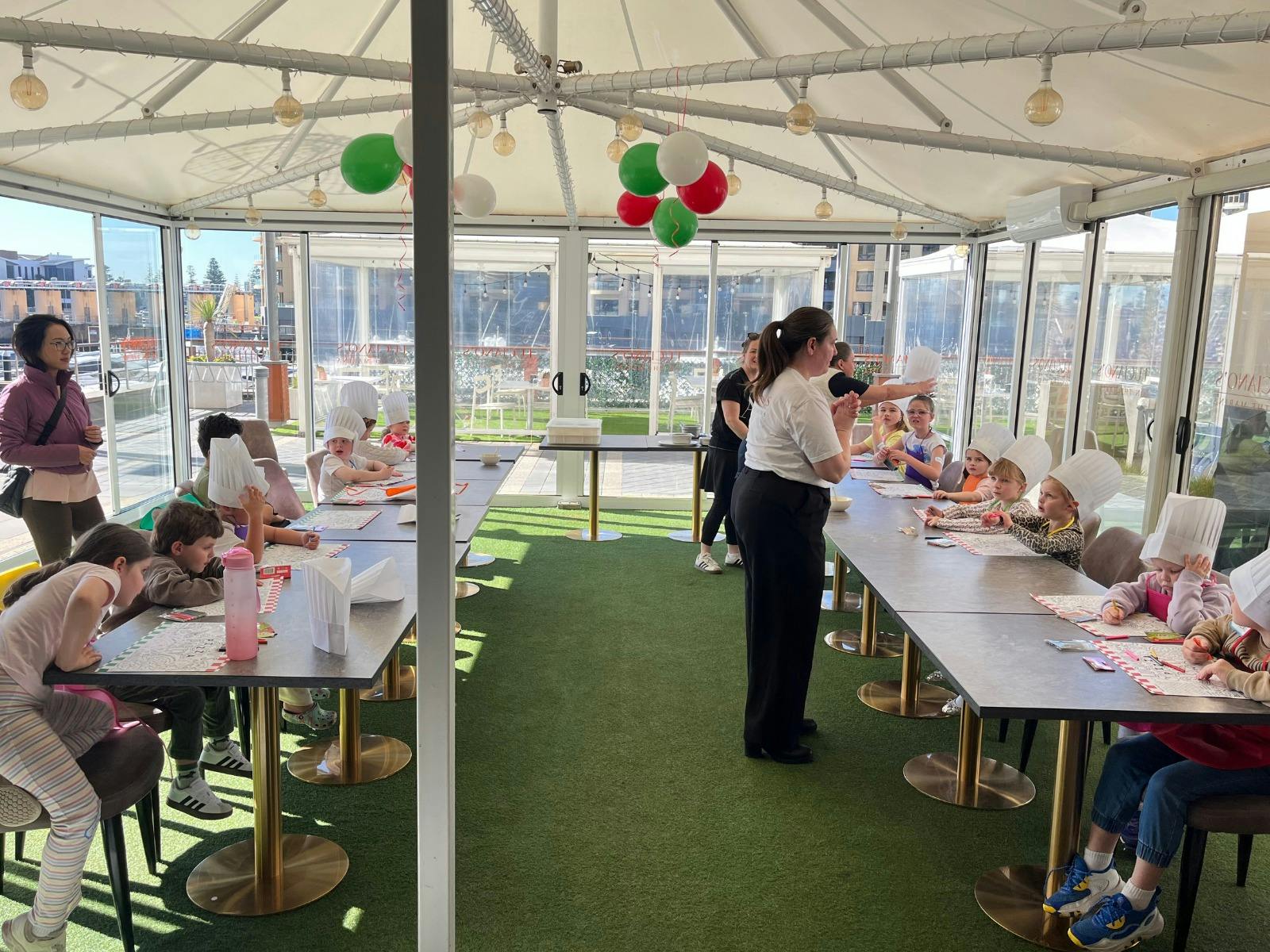 2 long tables with little kids sitting at them with chef hats on in a light filled room.