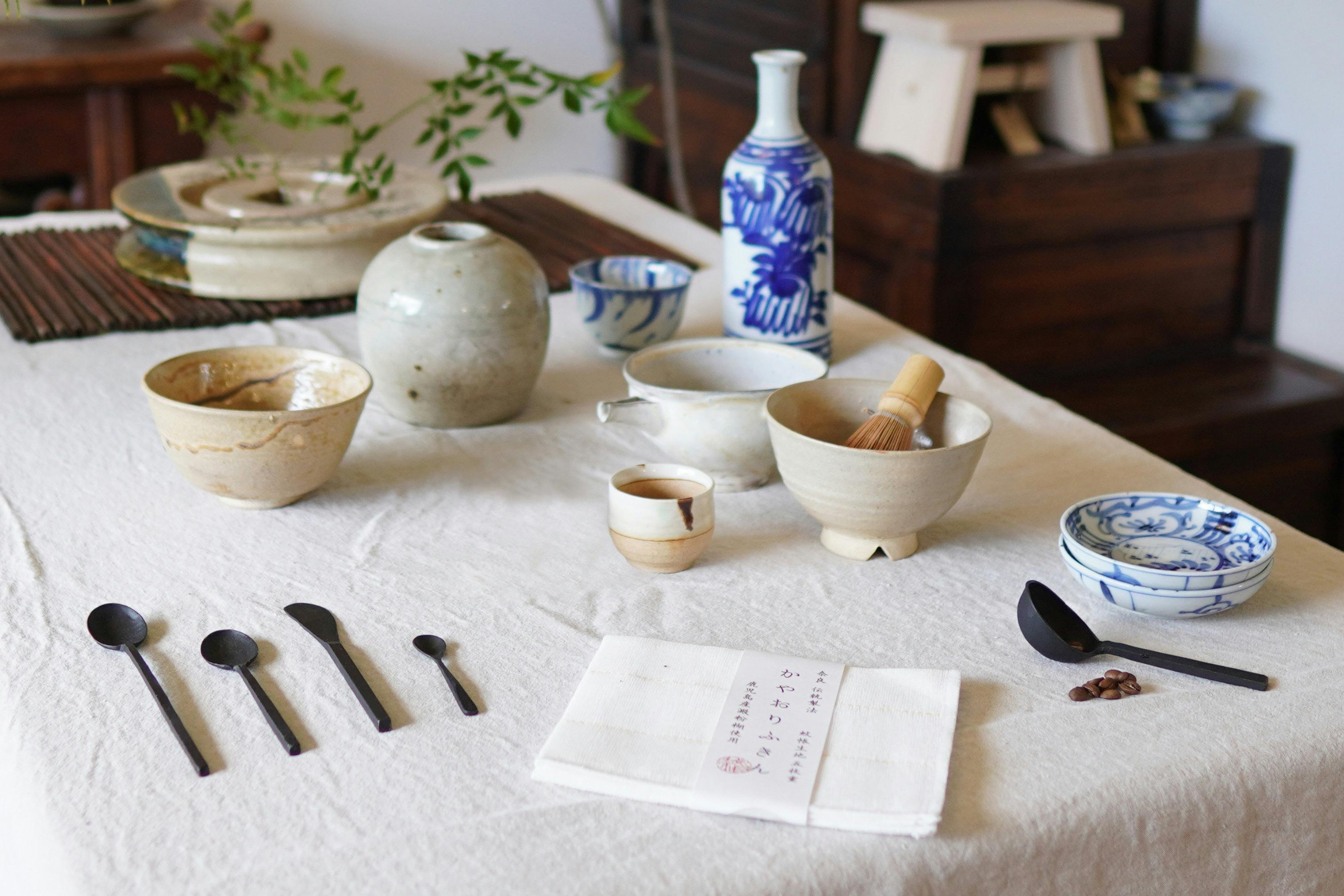 Photo of ceramic bowls, vases, dish and metal spoons on a table covered in linen