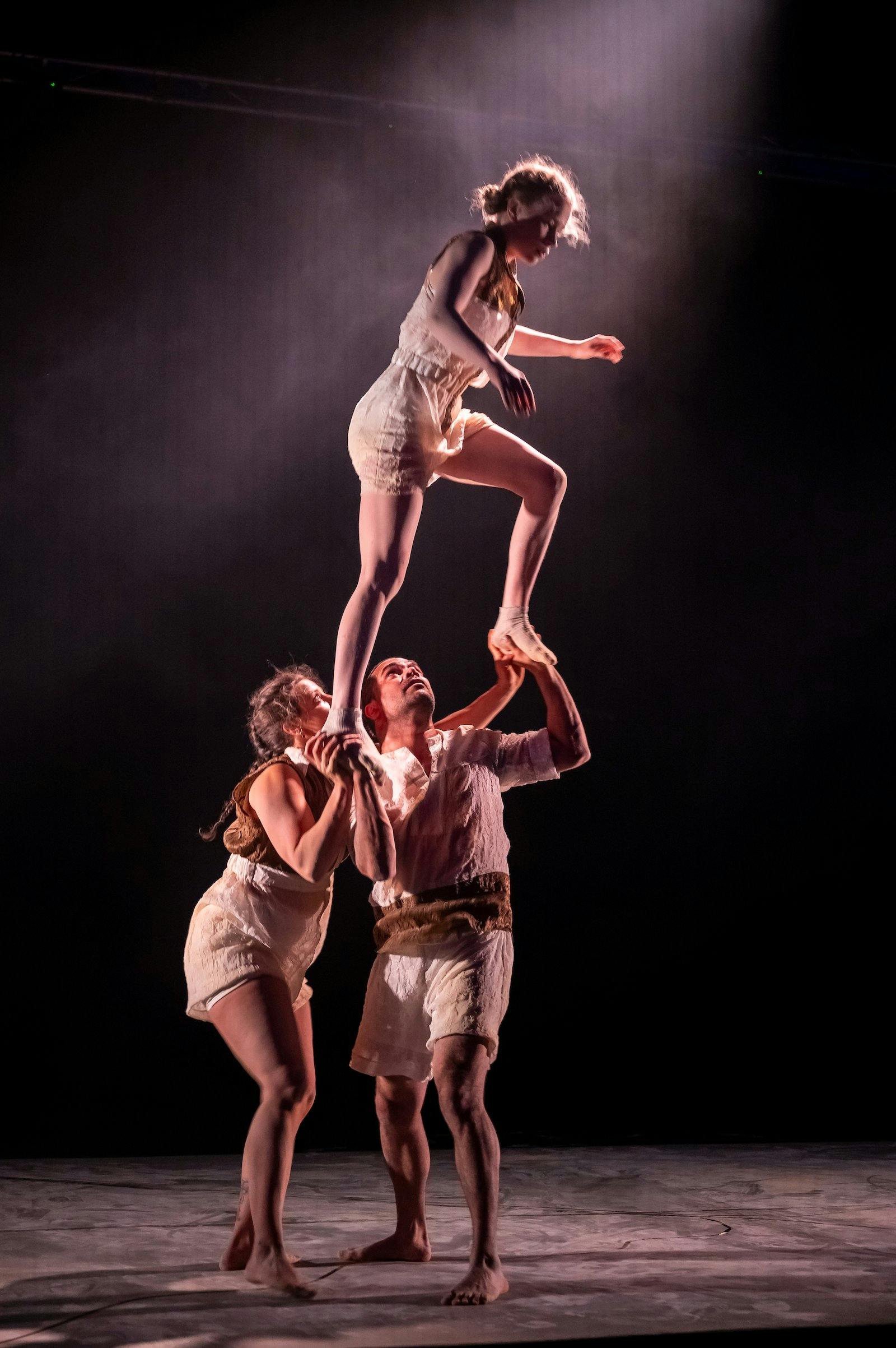 Three acrobats perform on stage under a spotlight, with two lifting a third person high. They wear l