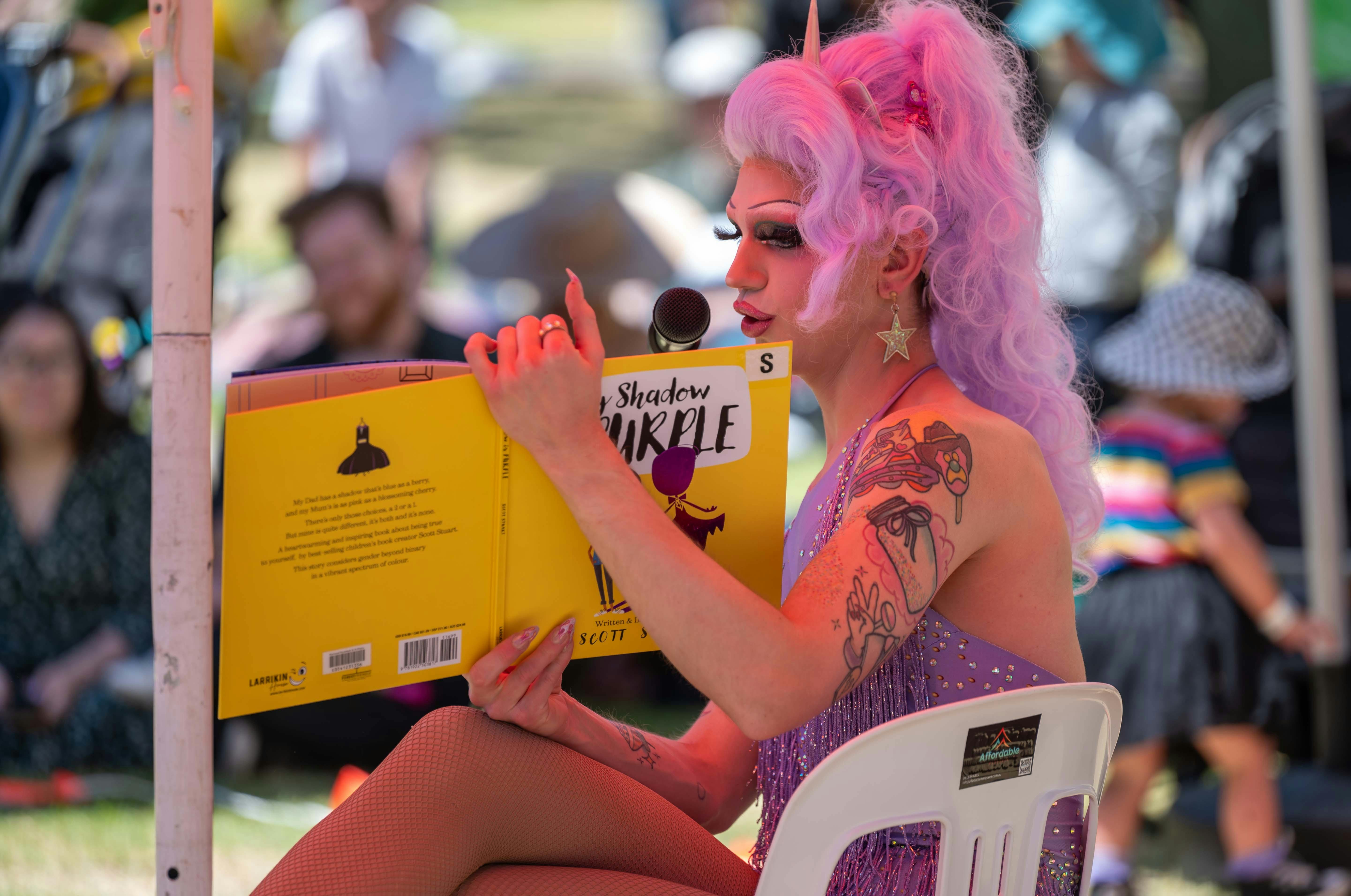 A drag performer with curly pastel purple hair, and a sparkly dress reads a book.