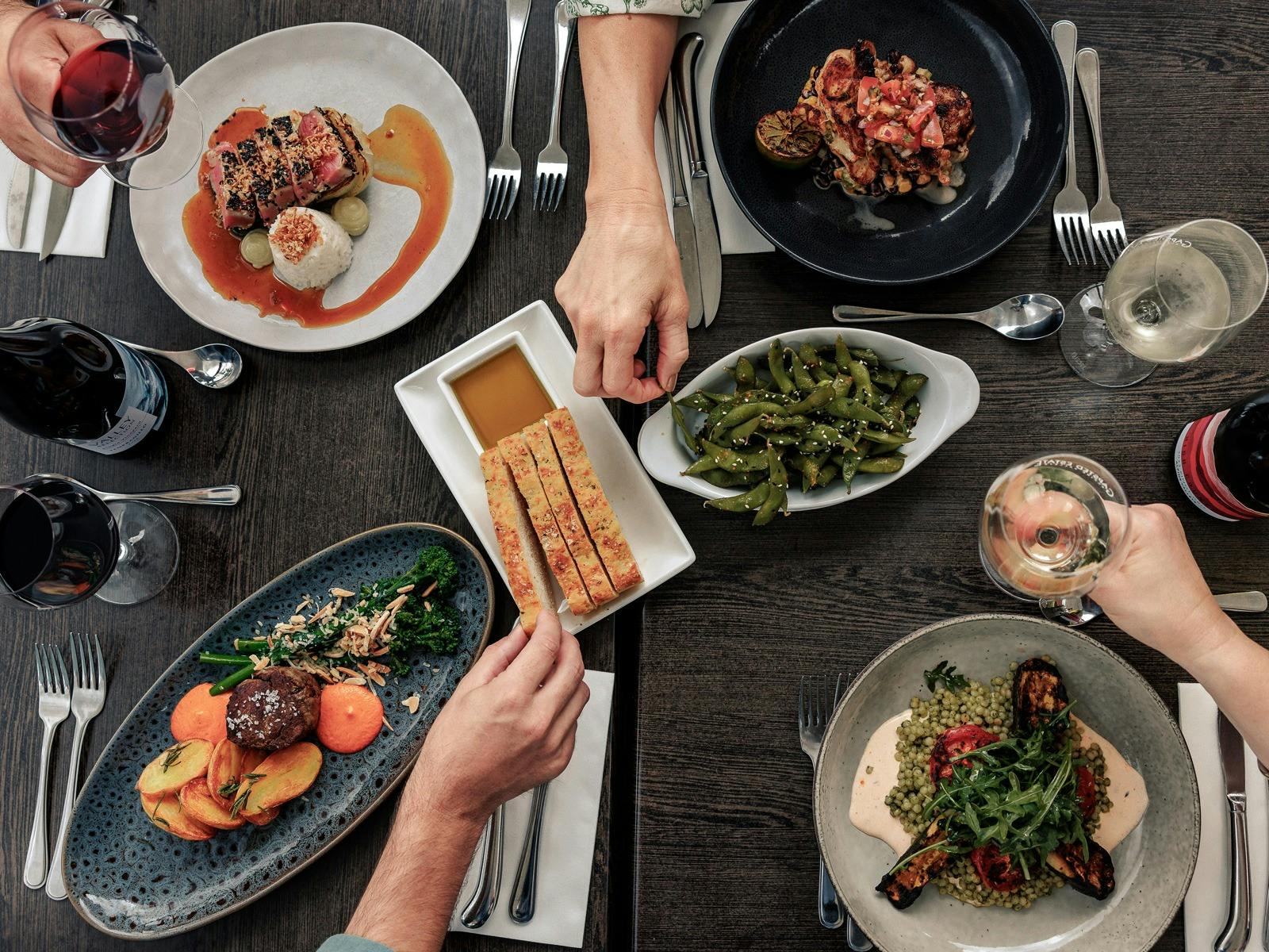 A table laid with seasonal dishes and wine, hands holding wine glasses and reaching for food