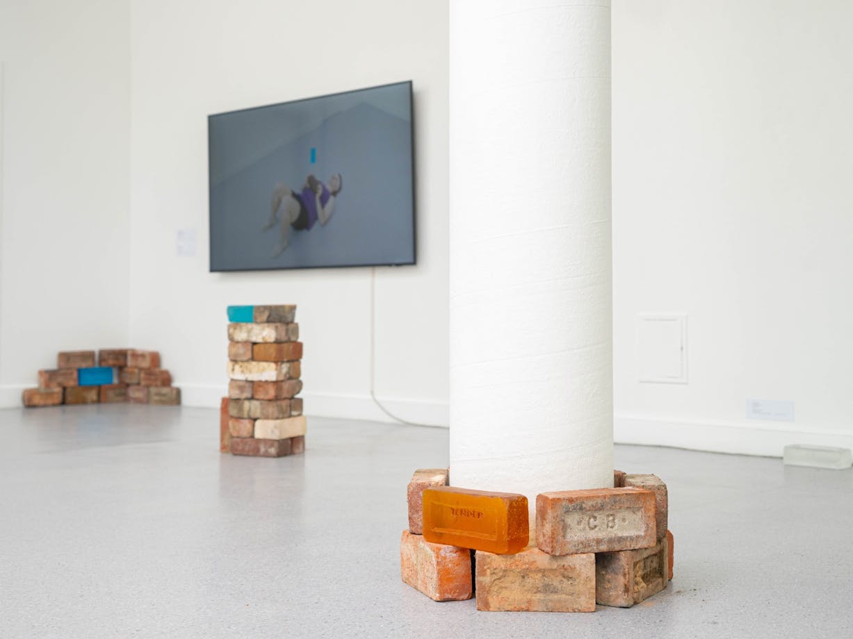 stacked glass and red bricks. TV instillation in the background featuring the artist lifting bricks.