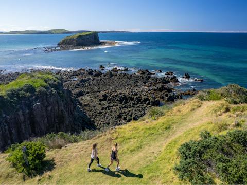 two girls walking on graddes headland with sun shining and blue water in background