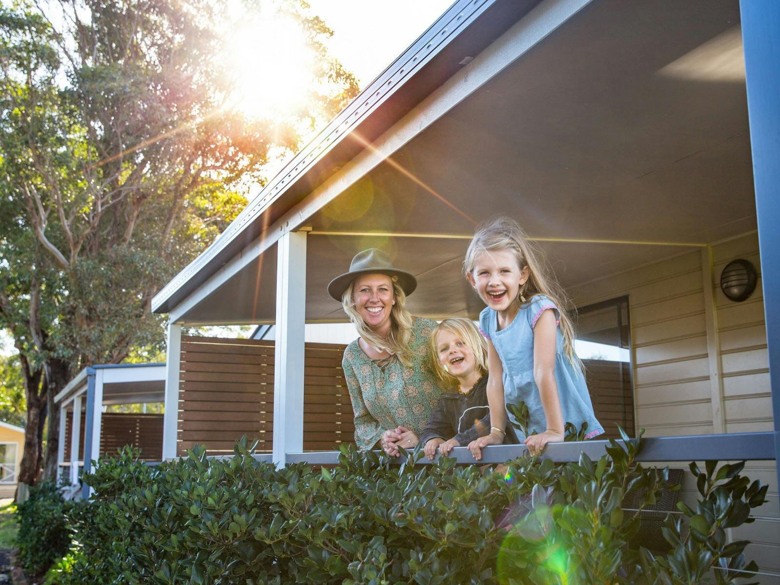 Mum and two young daughters smiling while they stand on the verandah of their cabin.