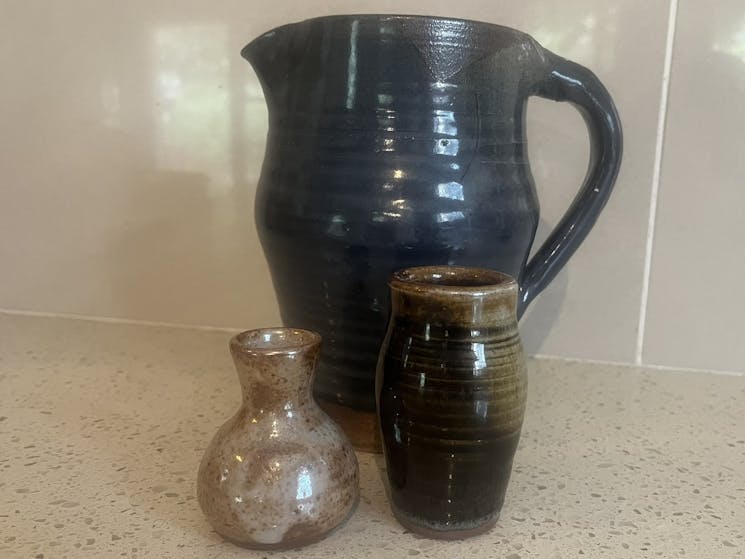 Three pots are against a cream wall.  Large black jug at the back, two smaller brown  pots in front