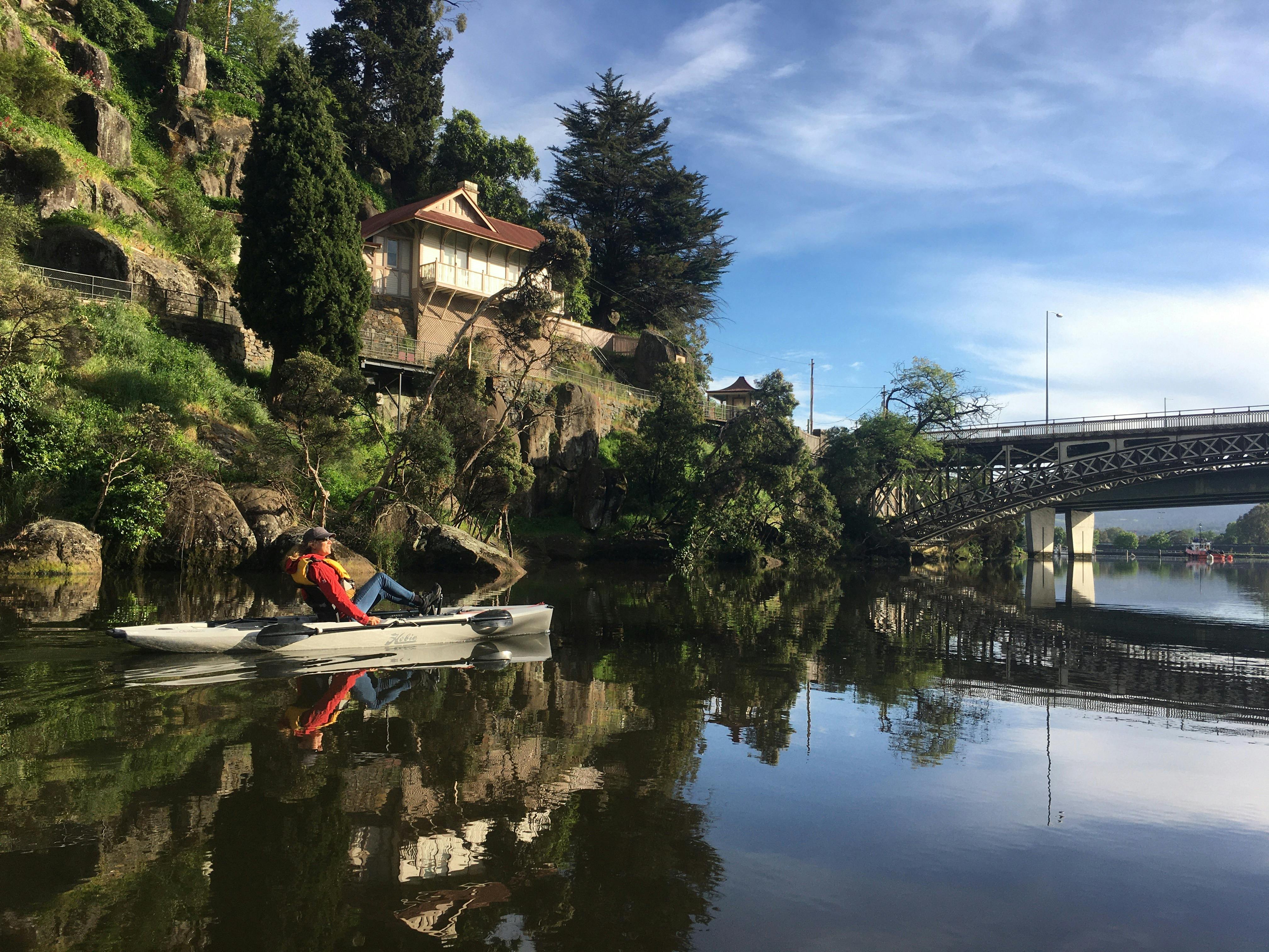 A woman in a red coat on a single pedal powered kayak passes a house overlooking reflective water