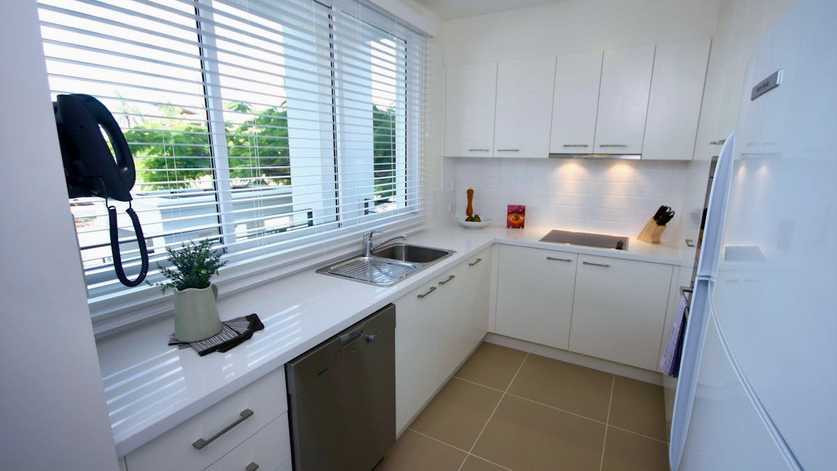 White modern kitchen with dishwasher, full-size refrigerator, stone benchtops and natural light thro