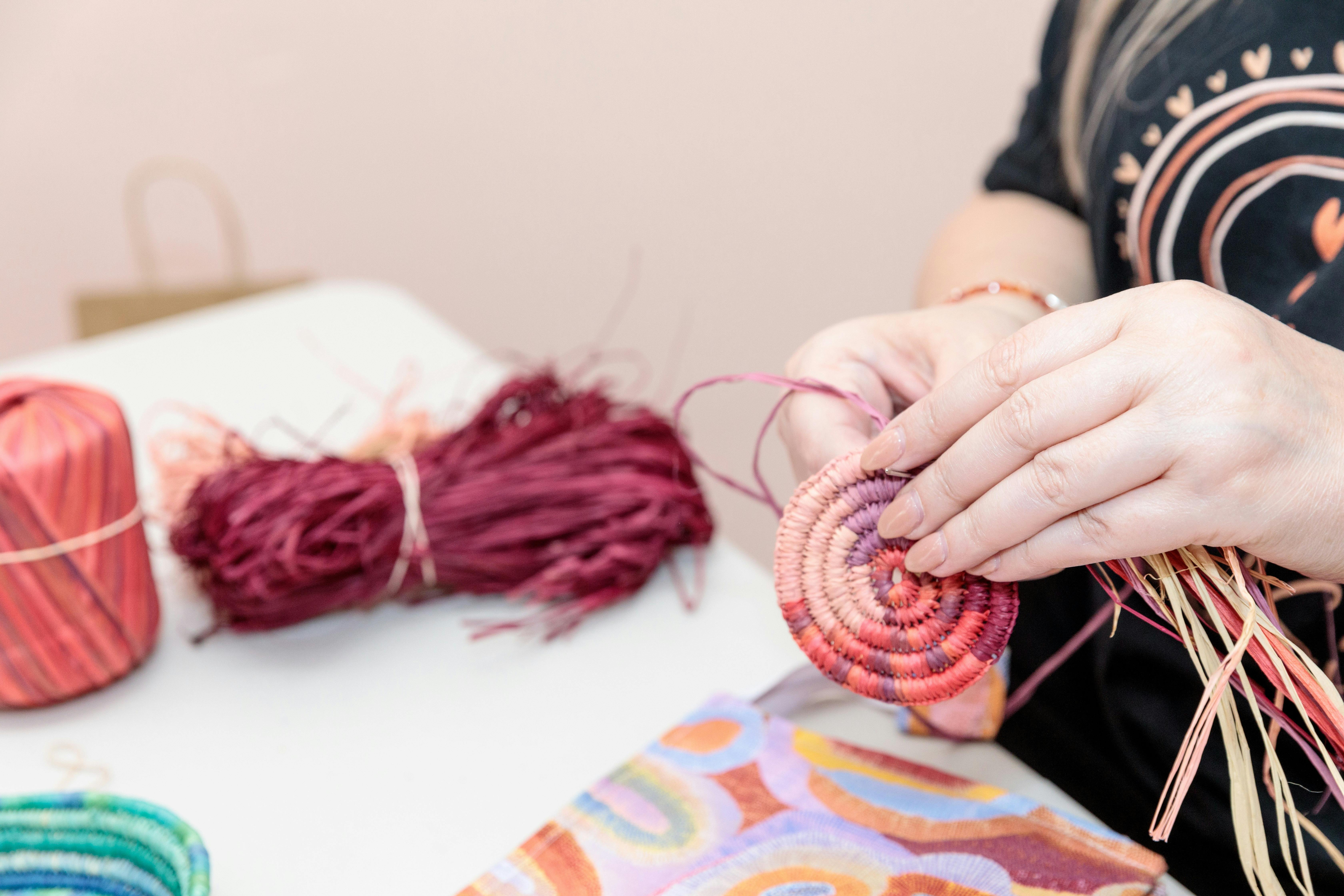 Picture shows a woman sitting at a table weavin g with red raffia