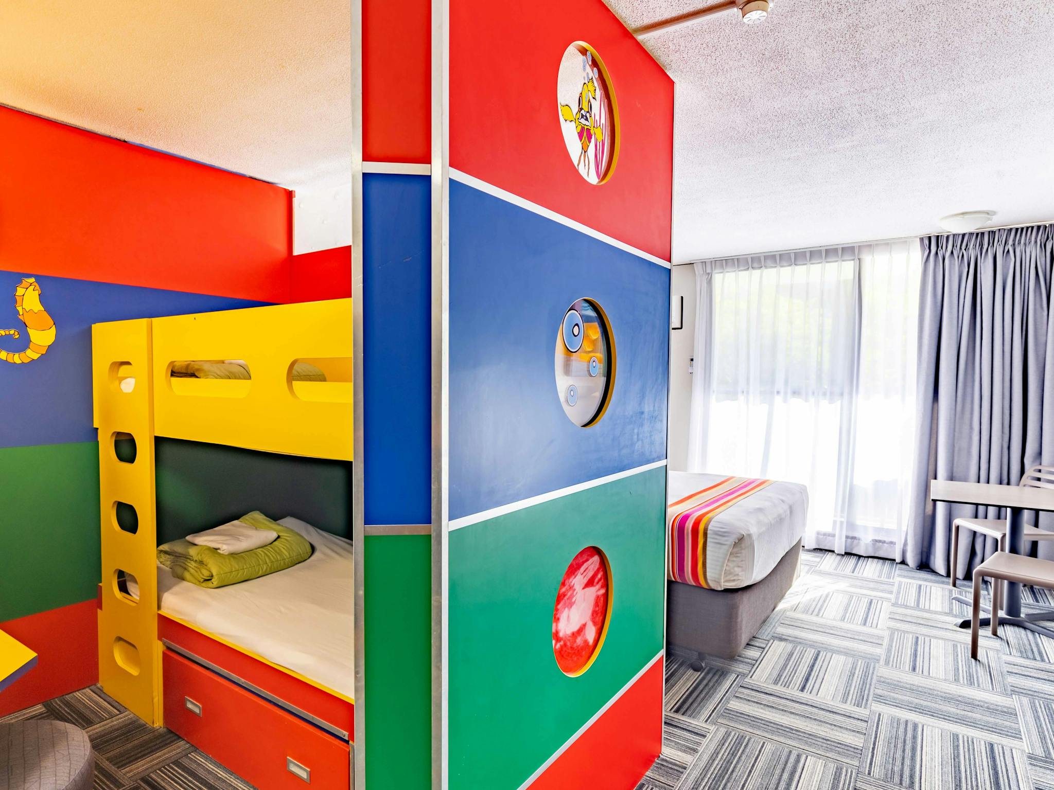 A room with a close image of the colourful set of bunk beds and the queen bed in the background A room with a close image of the colourful set of bunk beds and the queen bed in the background