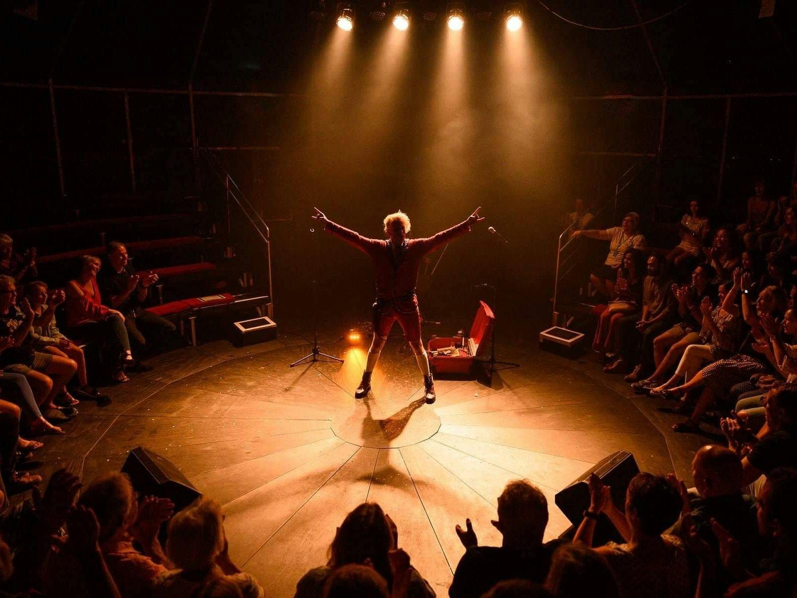 A lone performer poses ready for a comedy circus show, surrounded by a crowd on three sides.
