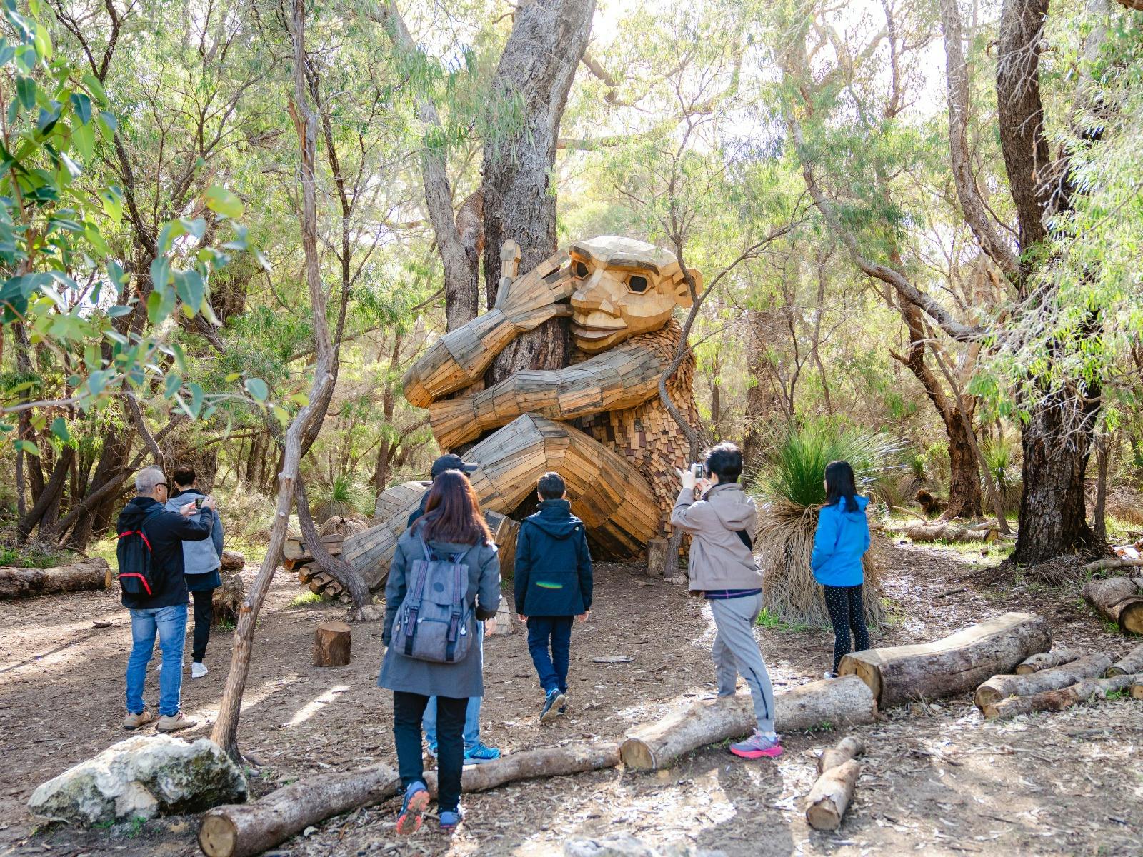 People gathered around a wooden 'troll-like' sculpture sat on the floor with its arms around a tree