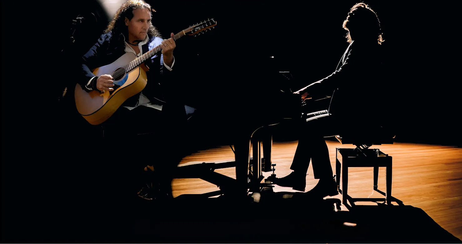 Two musicians performing on stage under low lighting: one playing an acoustic guitar and the other s