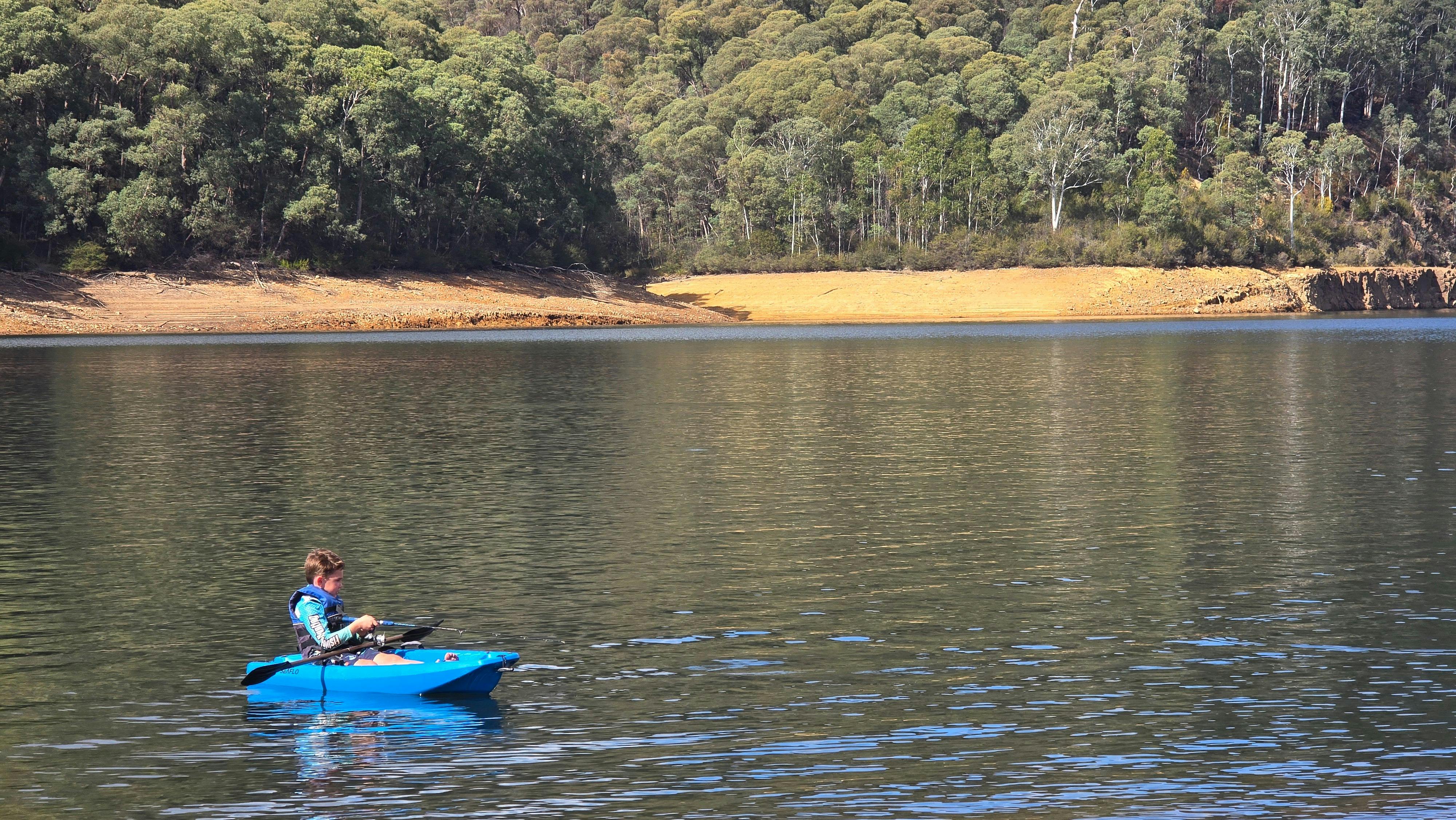 Young person fishing off a blue kayak on clear lake. Sand and trees in background.