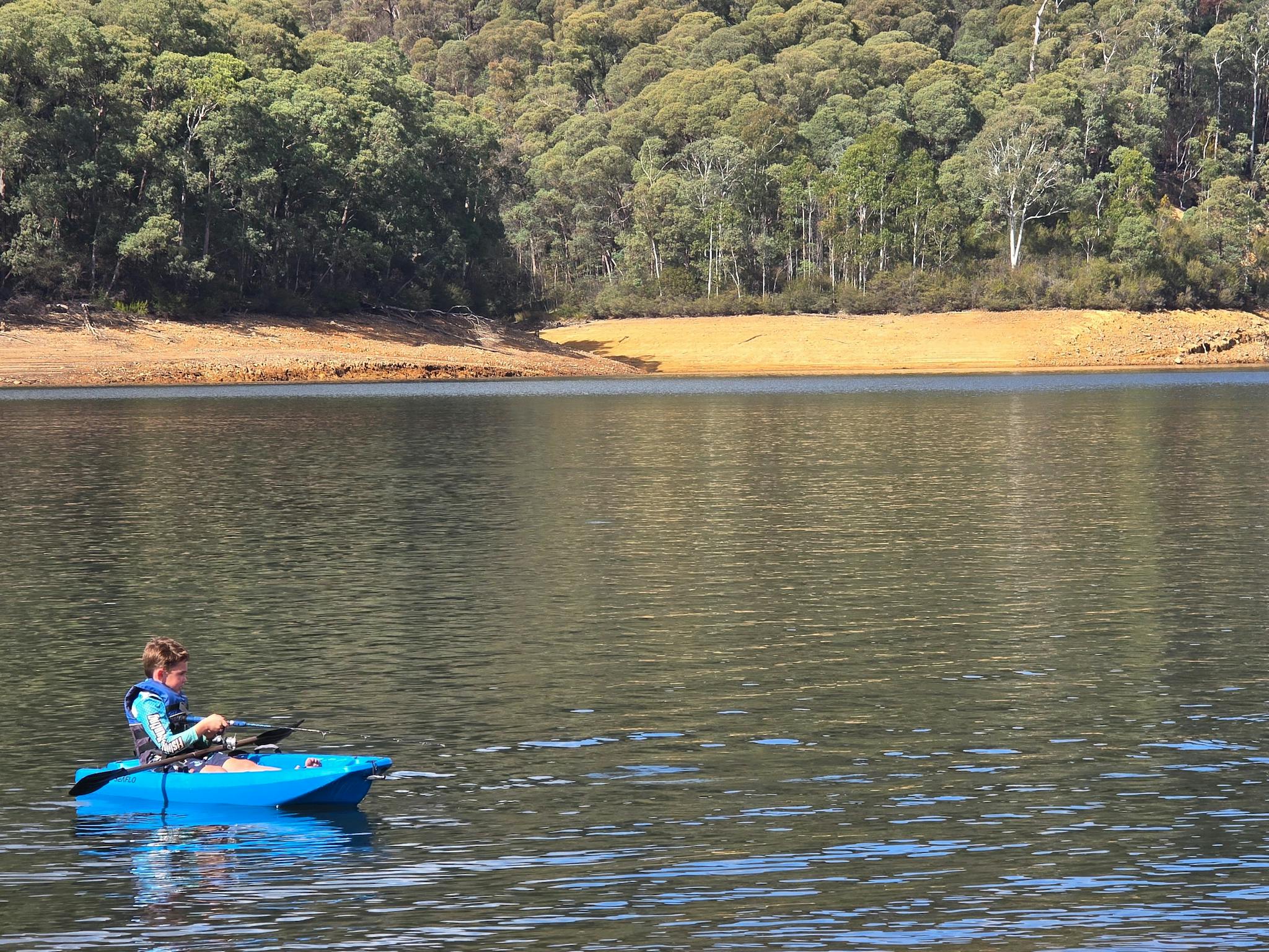 Young person fishing off a blue kayak on clear lake. Sand and trees in background.