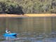 Young person fishing off a blue kayak on clear lake. Sand and trees in background.