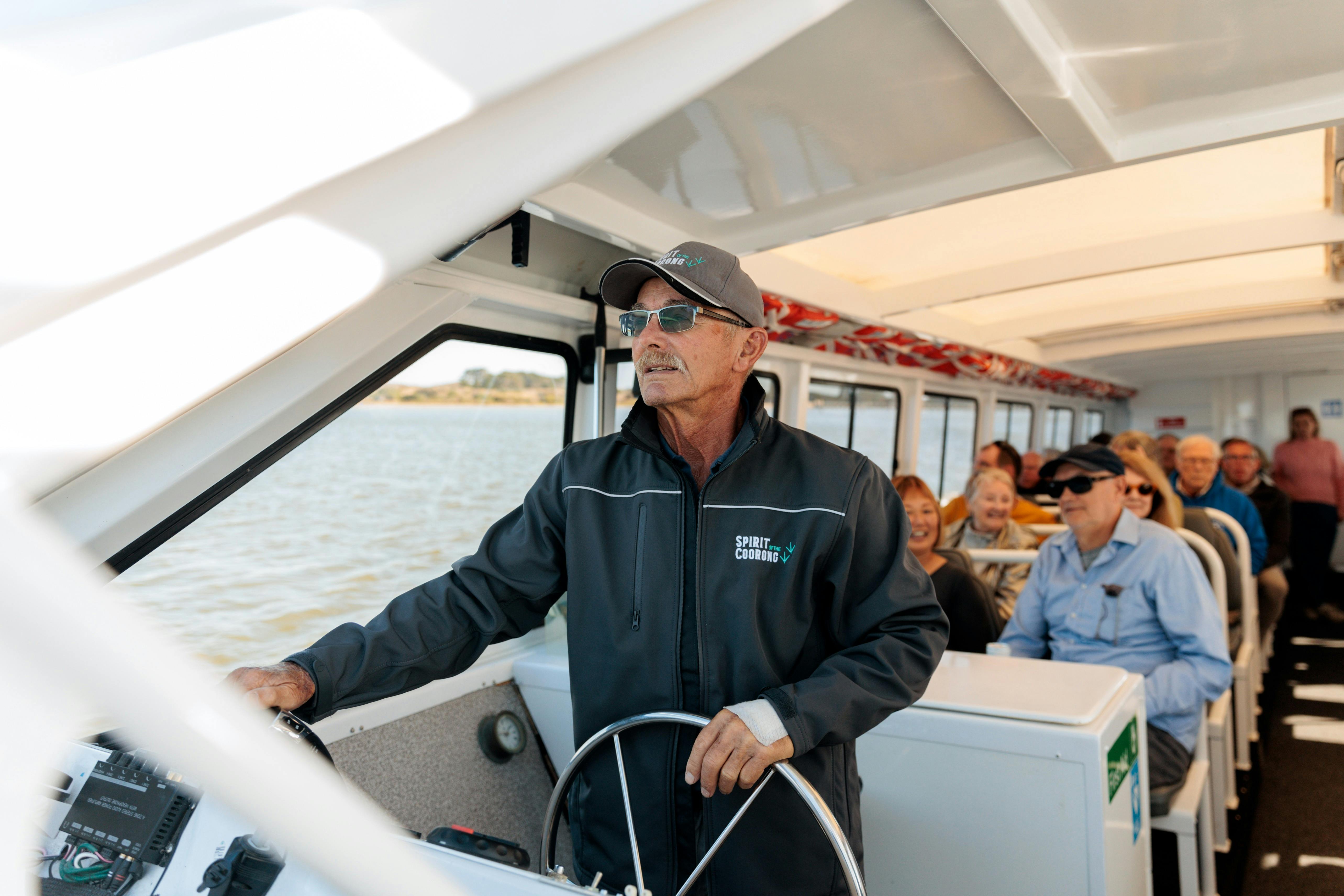 A captain steers the "Spirit of the Coorong" boat, with smiling passengers seated behind him.