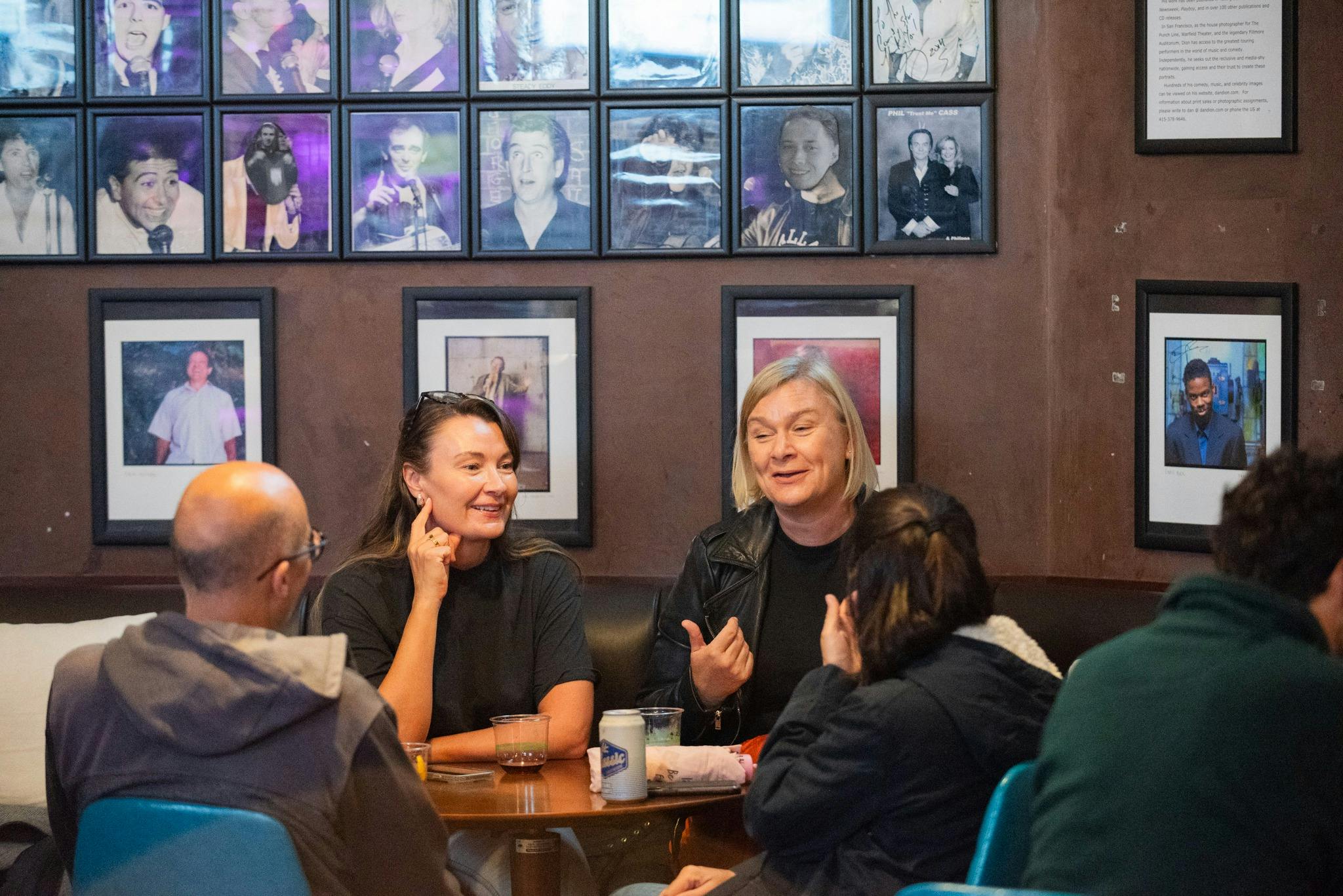 A group of people sitting and talking in a cafe with framed images of comedians on the wall behind