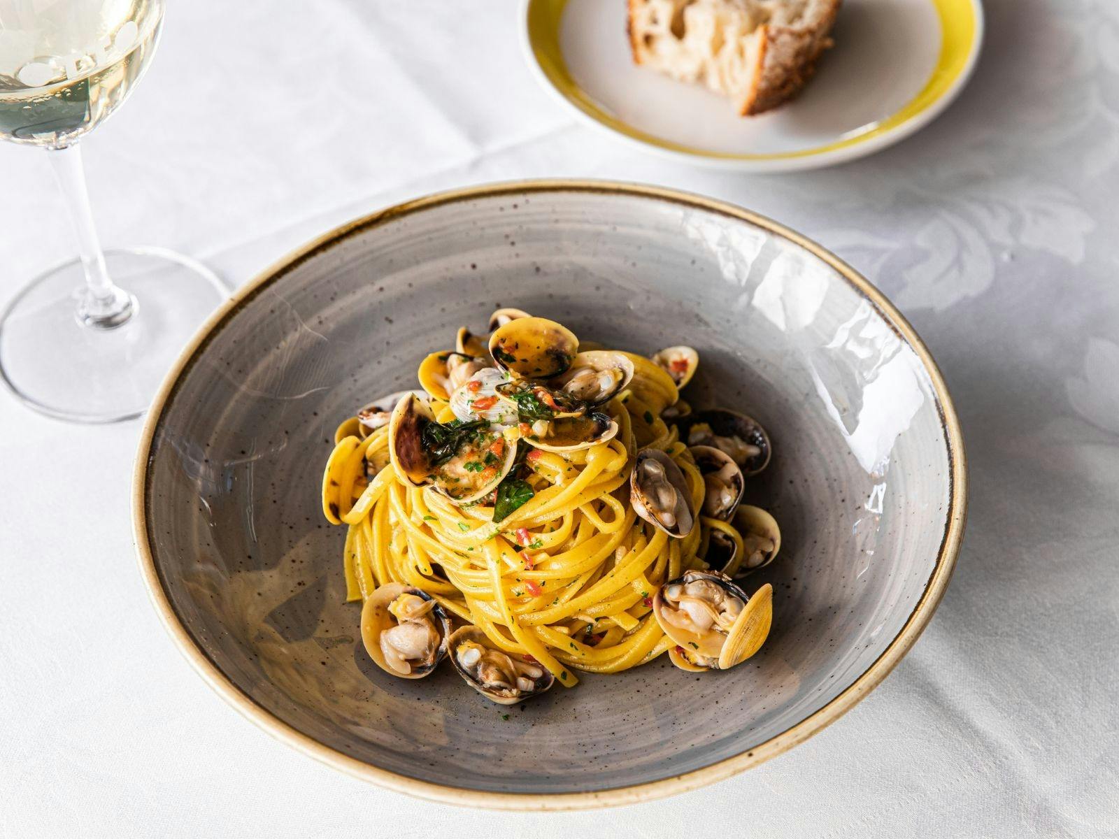 Bowl of spaghetti e vongole on a white table cloth, with a glass of white wine and bread slice.