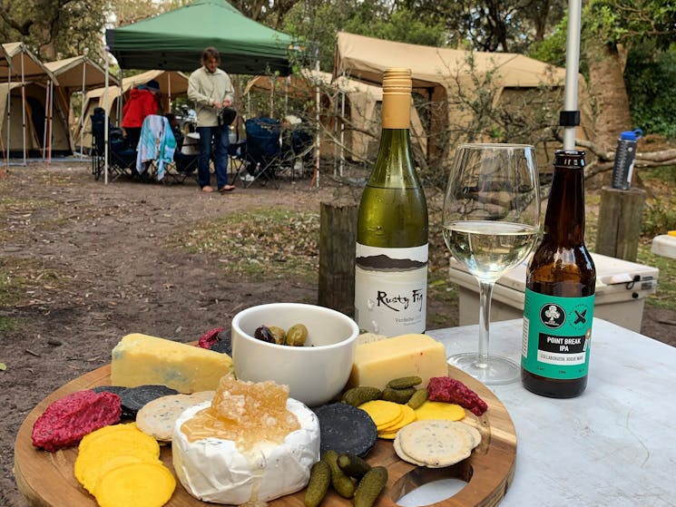 A cheese board in the foreground with tents and campsite behind