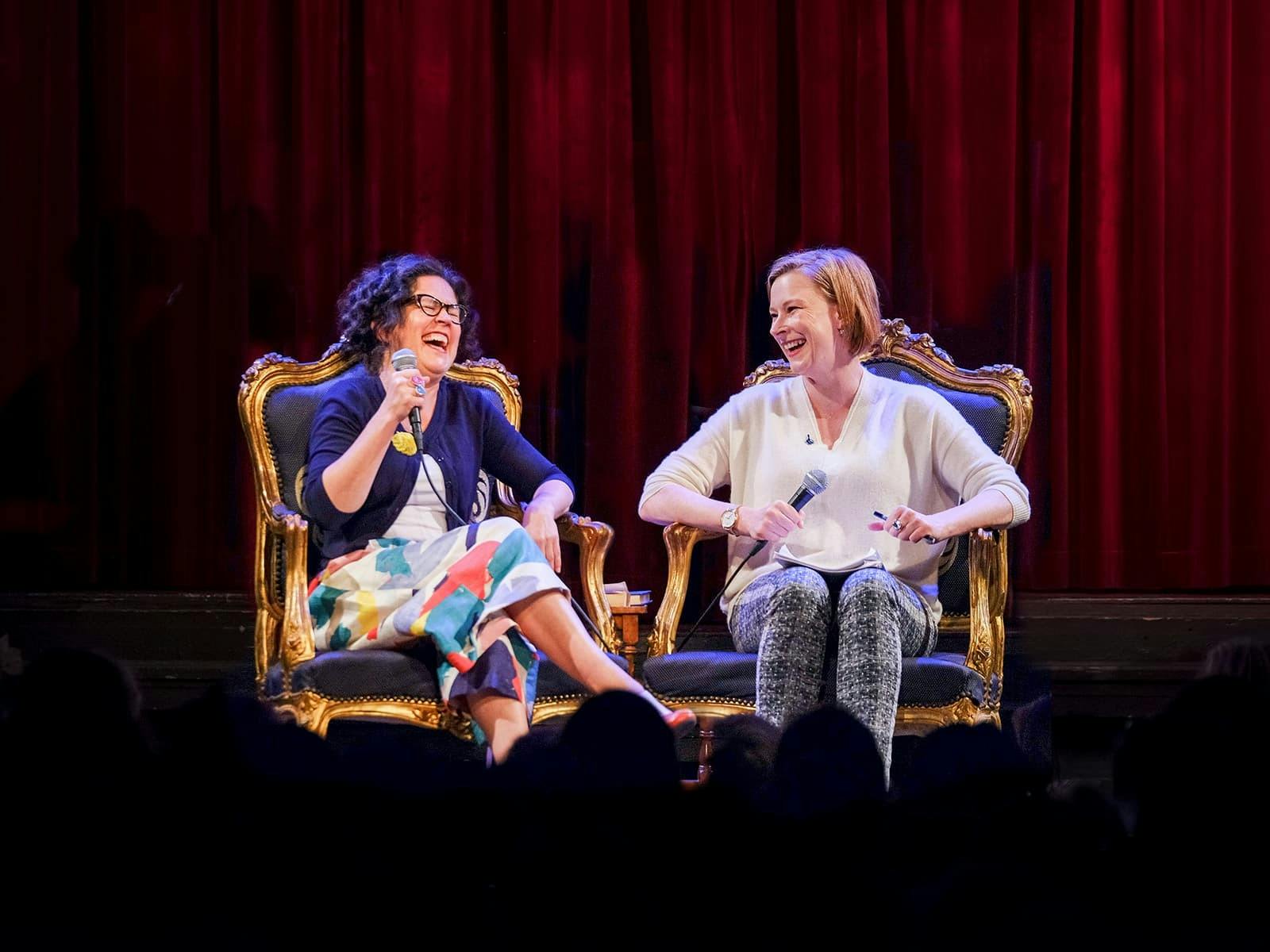 Annabel Crabb & Leigh Sales sitting in lush chairs on stage, both are holding microphones & laughing