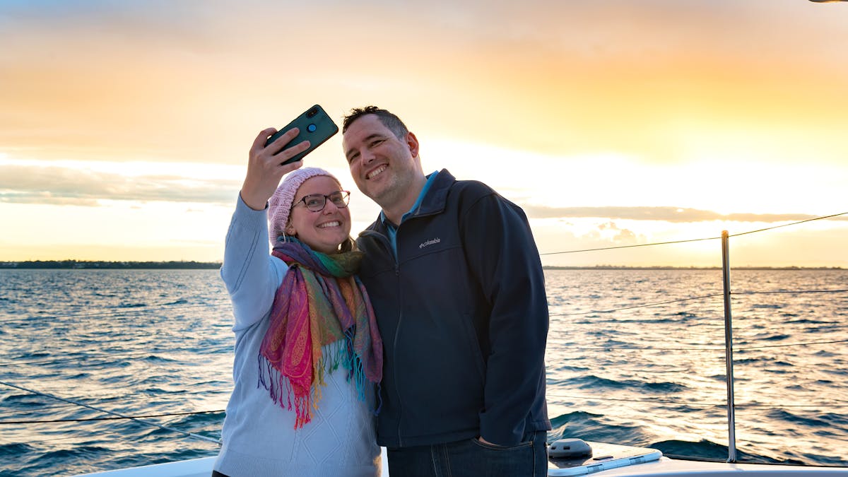 Couple taking a selfie on a boat as sun sets over the water