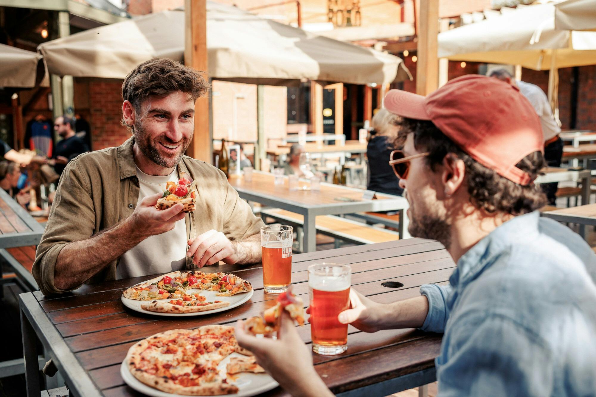Two men eating woodfired pizza and drinking pints of beer in a warmly lit beer garden