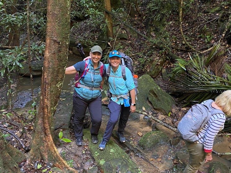 Two women dressed in blue pose near a tree on a hill with backpacks and hiking gear on