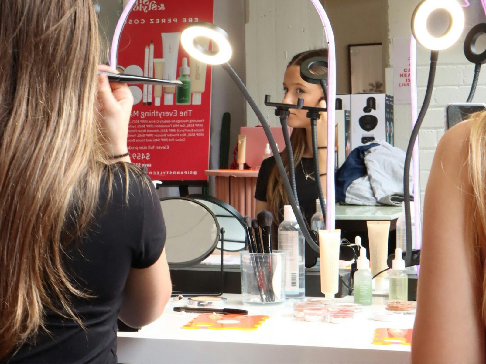 Woman applying makeup at a lit vanity with products, brushes, and her reflection in the mirror.