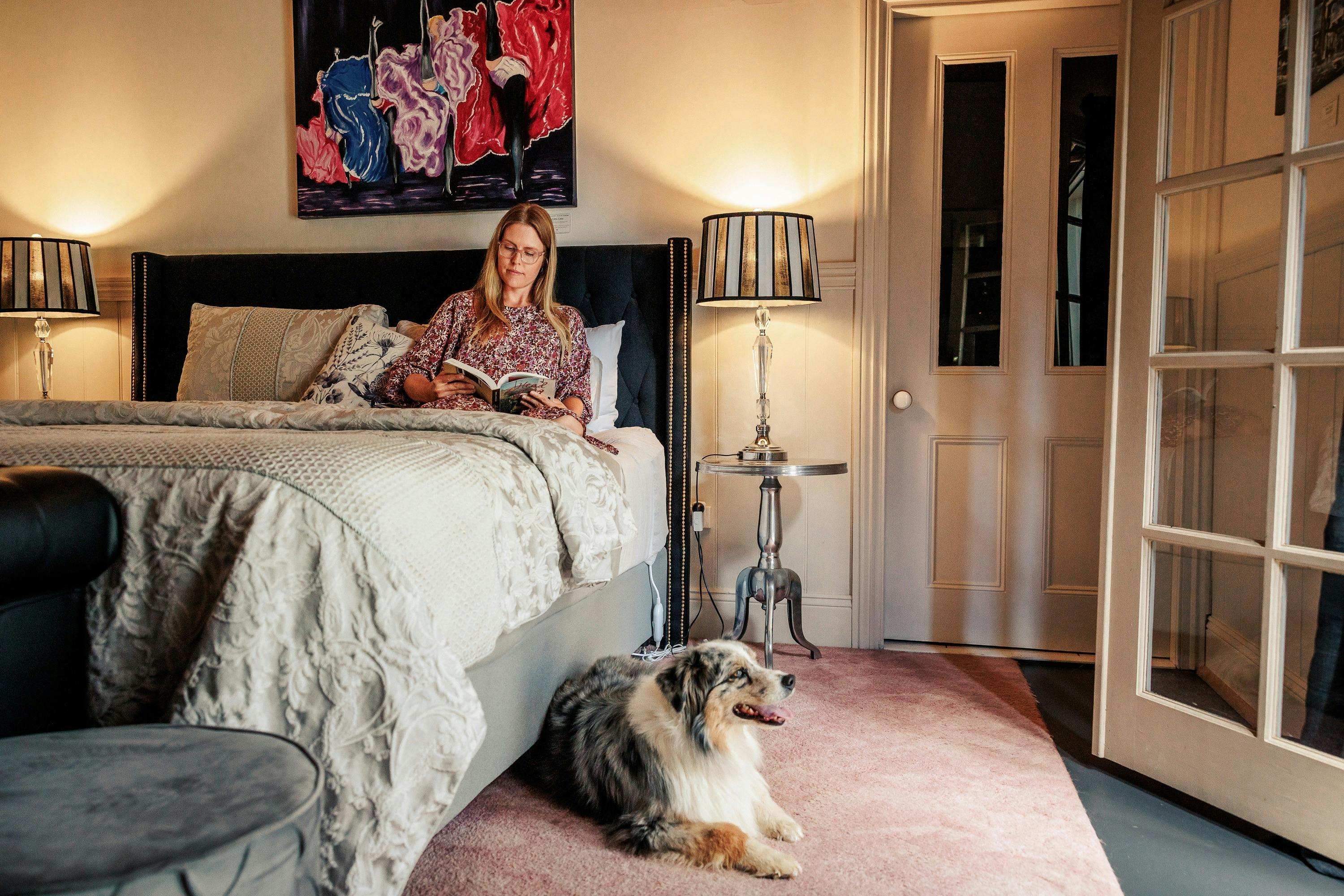 Woman reading while sitting on the bed with a dog seated by the bed, table lamps lit