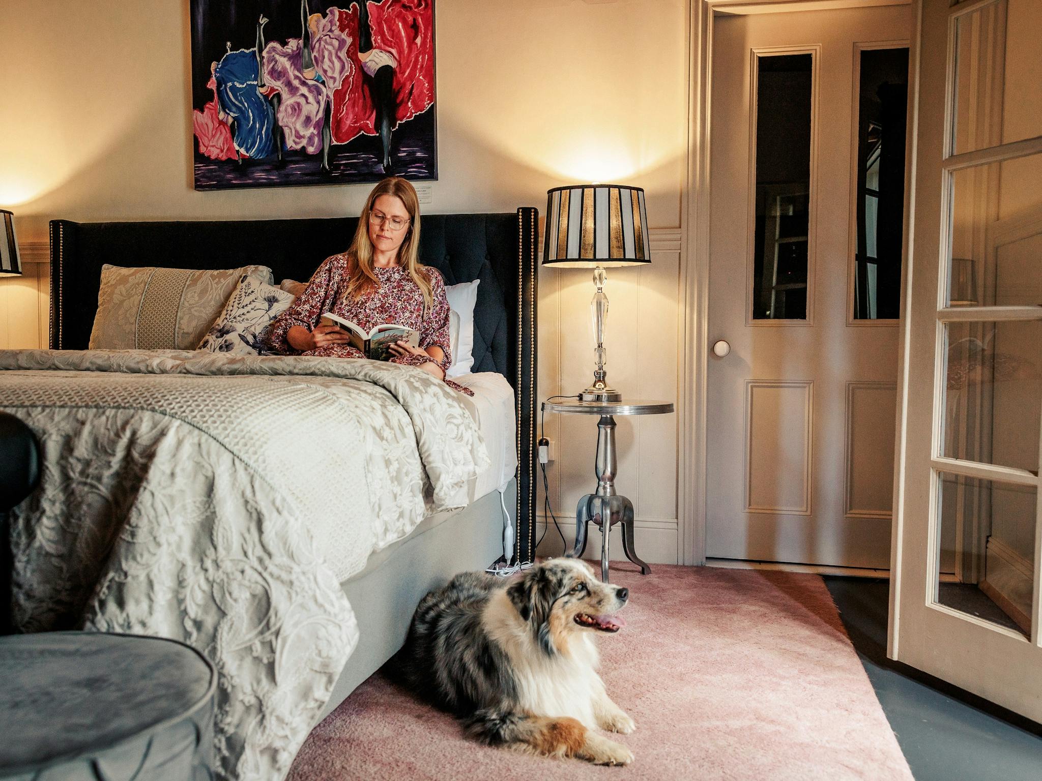Woman reading while sitting on the bed with a dog seated by the bed, table lamps lit