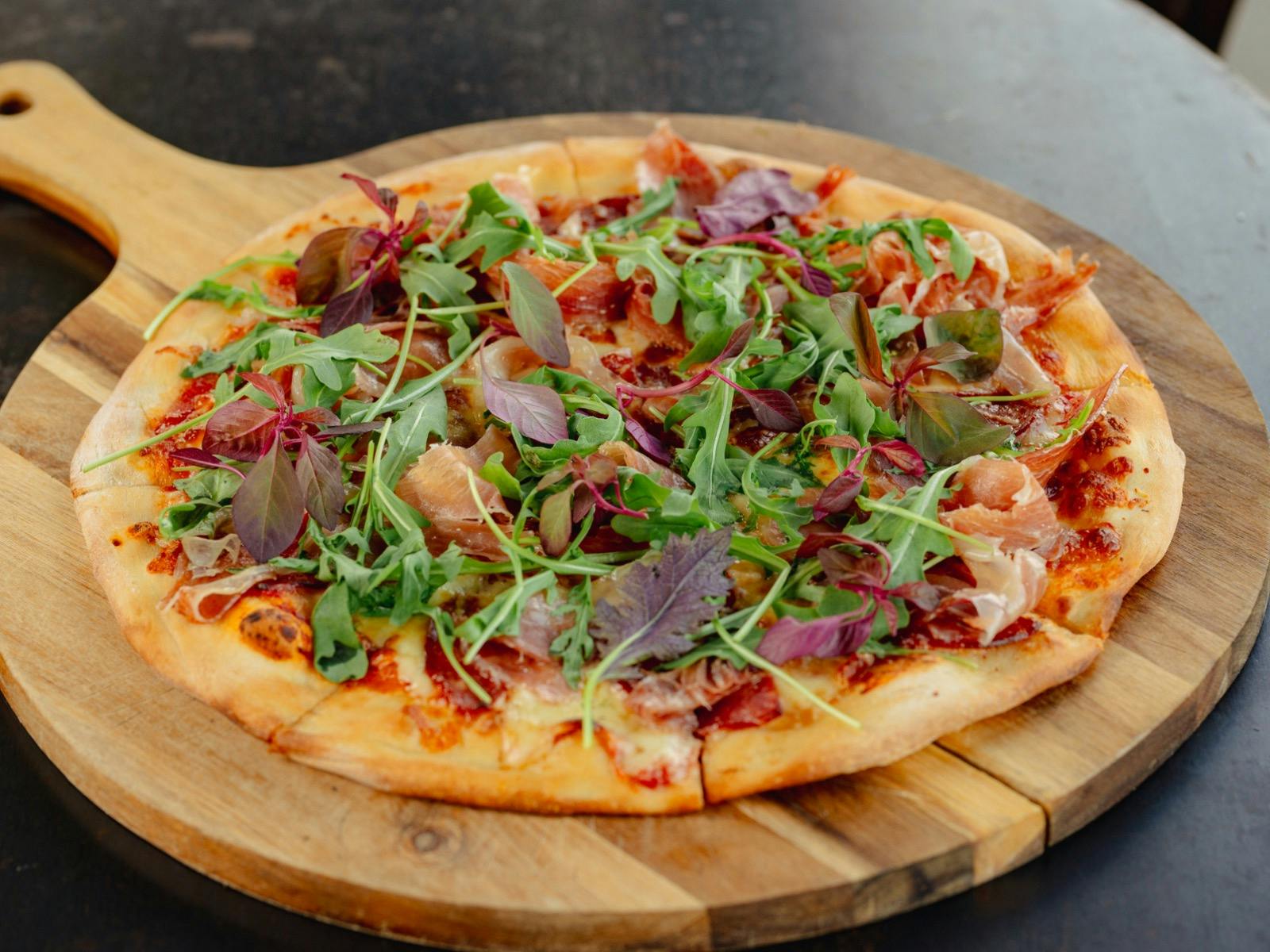 A large pizza on a wooden board topped with arugula, prosciutto and mozzarella.