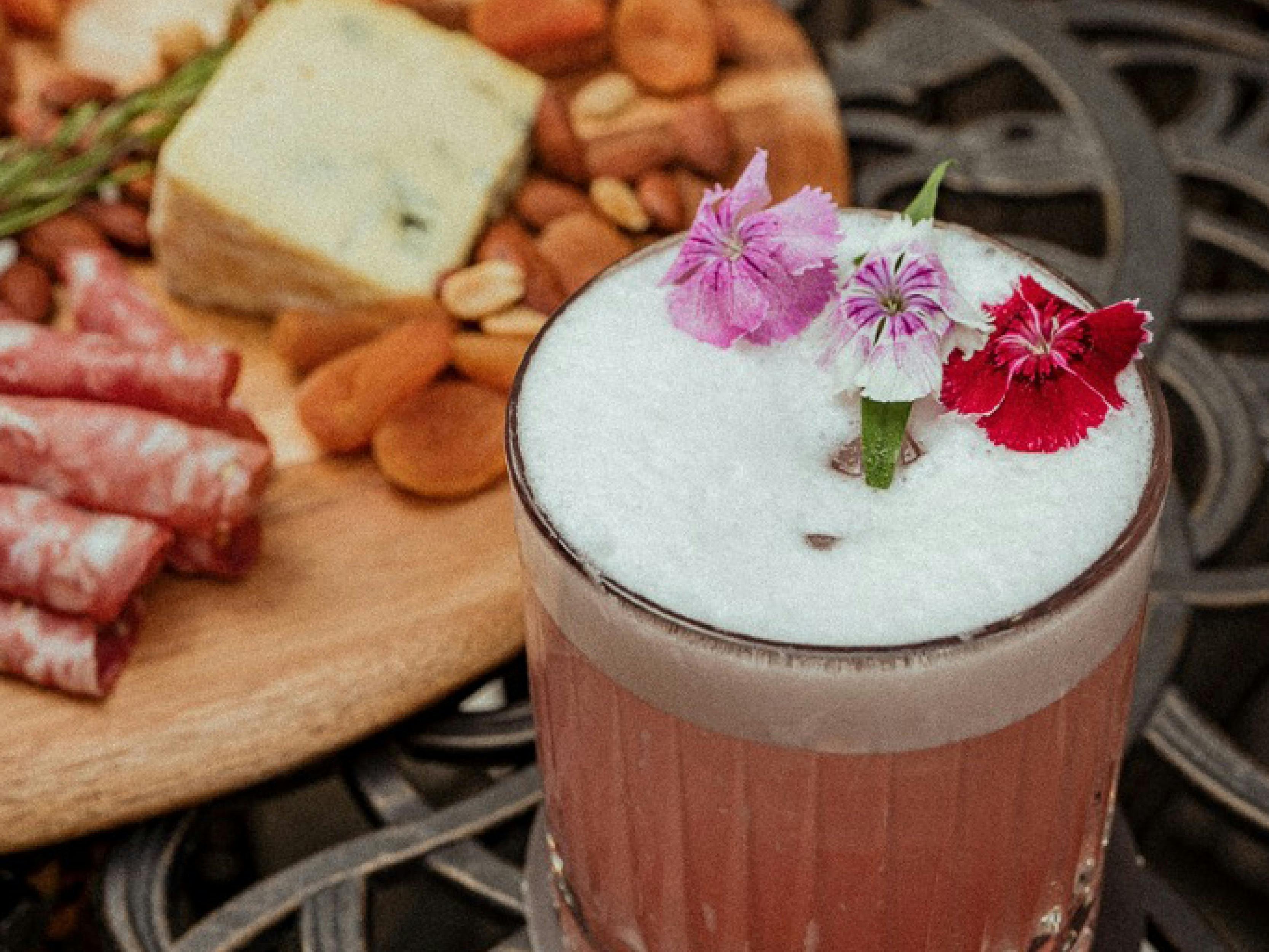 A photo of a cocktail with a edible flower garnish, in front of a cheeseboard.