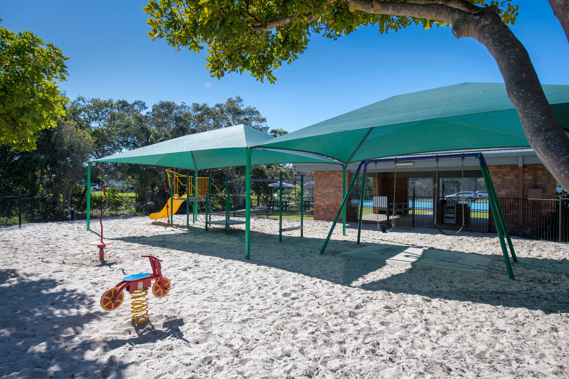 Dicky Beach Holiday Park playground on sand with shade covers and tennis court in background