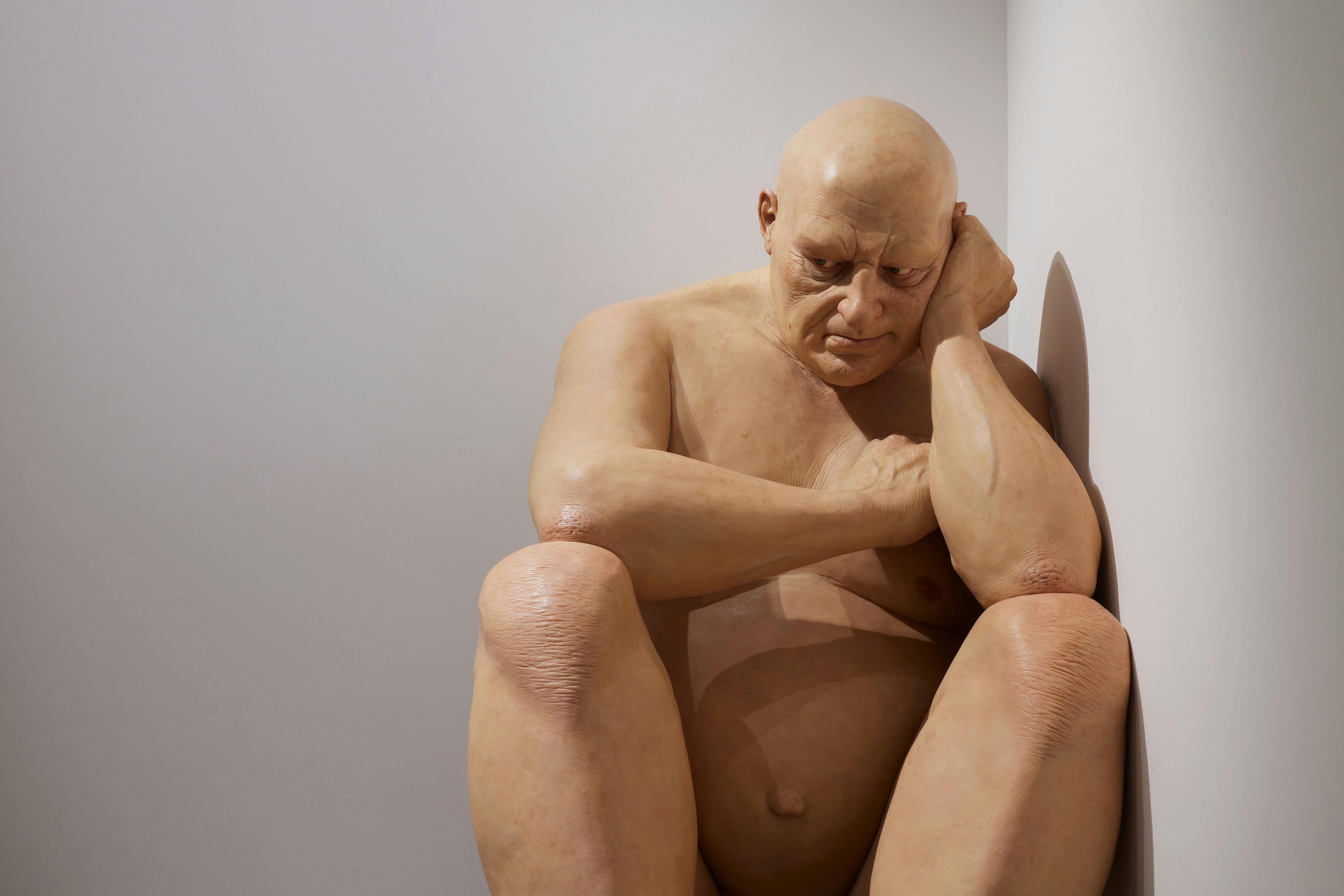 a large hyper realistic sculpture of a grump old man slumped in the corner
