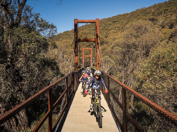 A family bike rides across a suspension bridge along Thredbo Valley track, Kosciuszko National Park