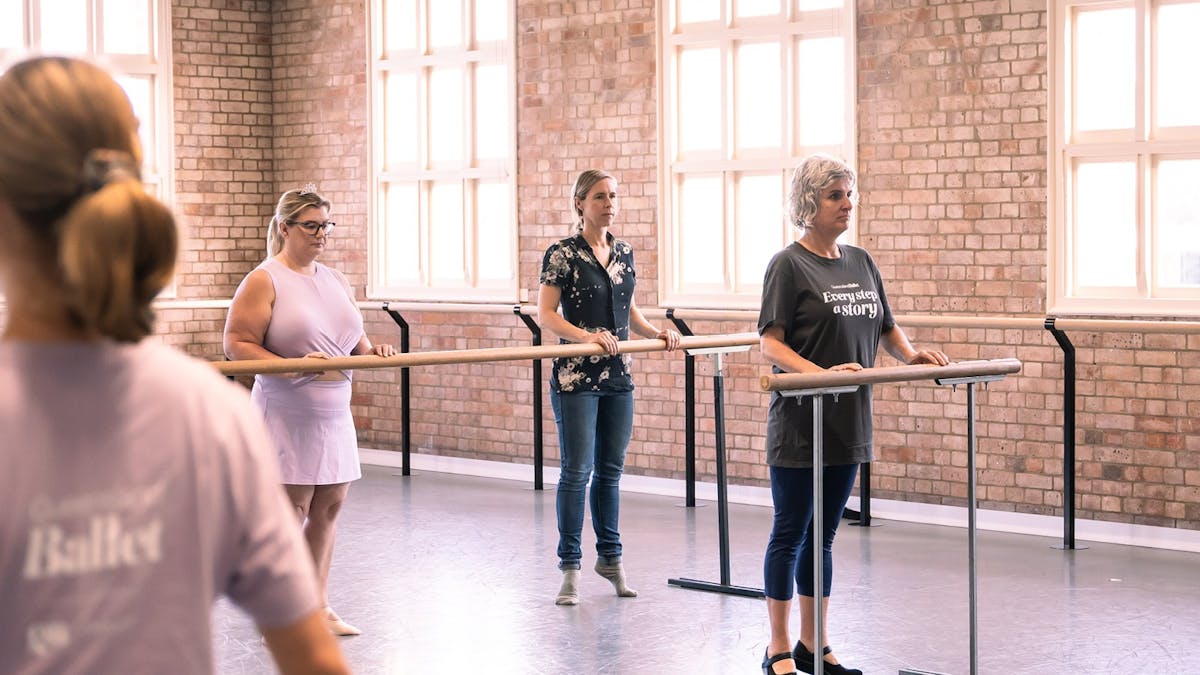 3 ladies holding onto ballet barre on raised toes under the guidance of a Queensland Ballet teacher