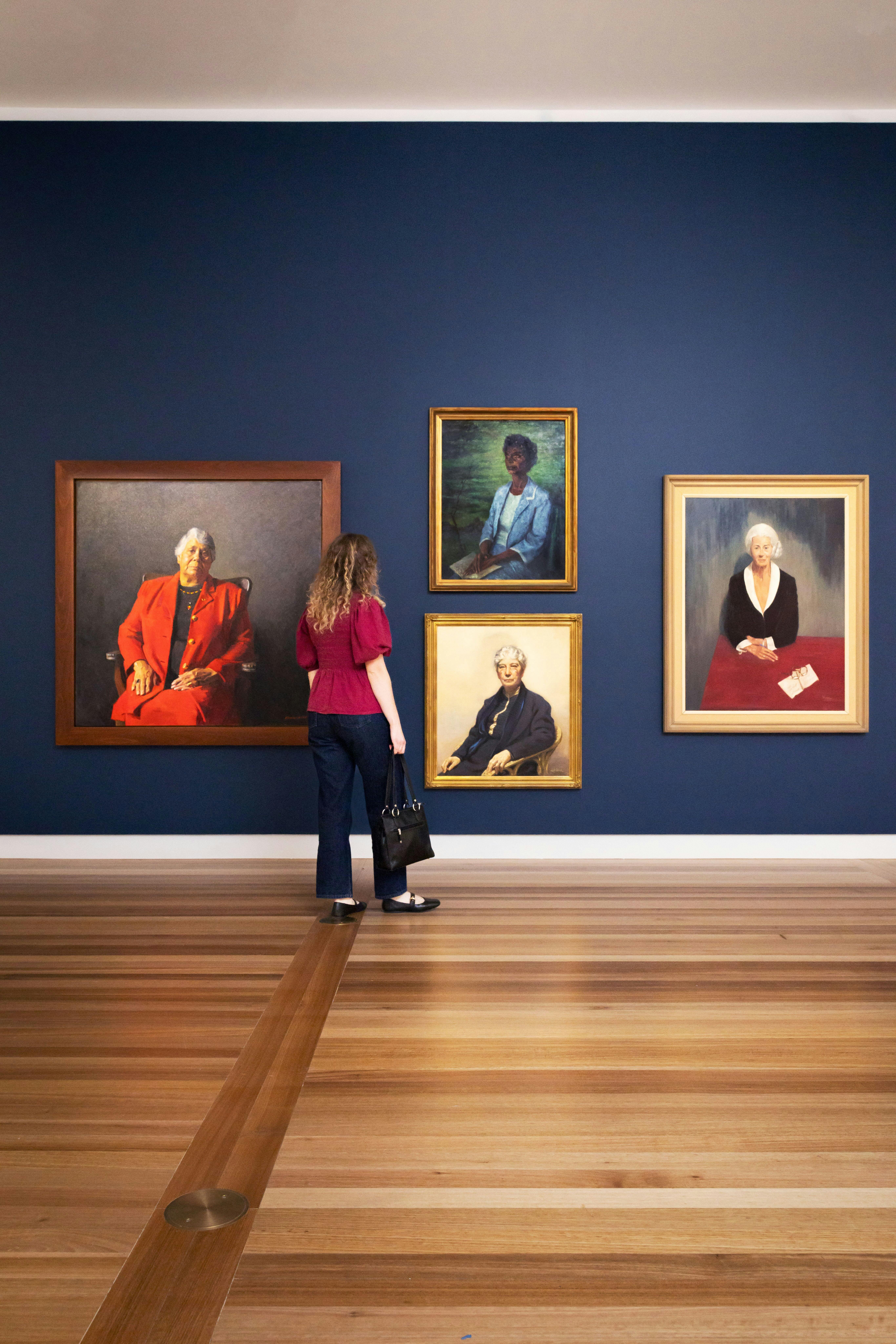 A woman looks at a dark wall featuring four large painted portraits of various women wearing suits.