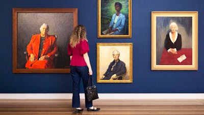 A woman looks at a dark wall featuring four large painted portraits of various women wearing suits.