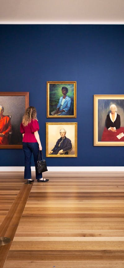 A woman looks at a dark wall featuring four large painted portraits of various women wearing suits.