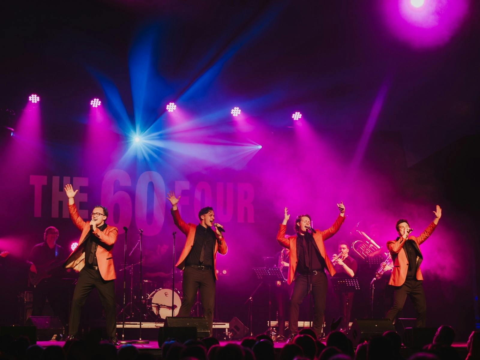 Band performs energetically on stage in orange jackets, with vibrant lights and a live band behind.