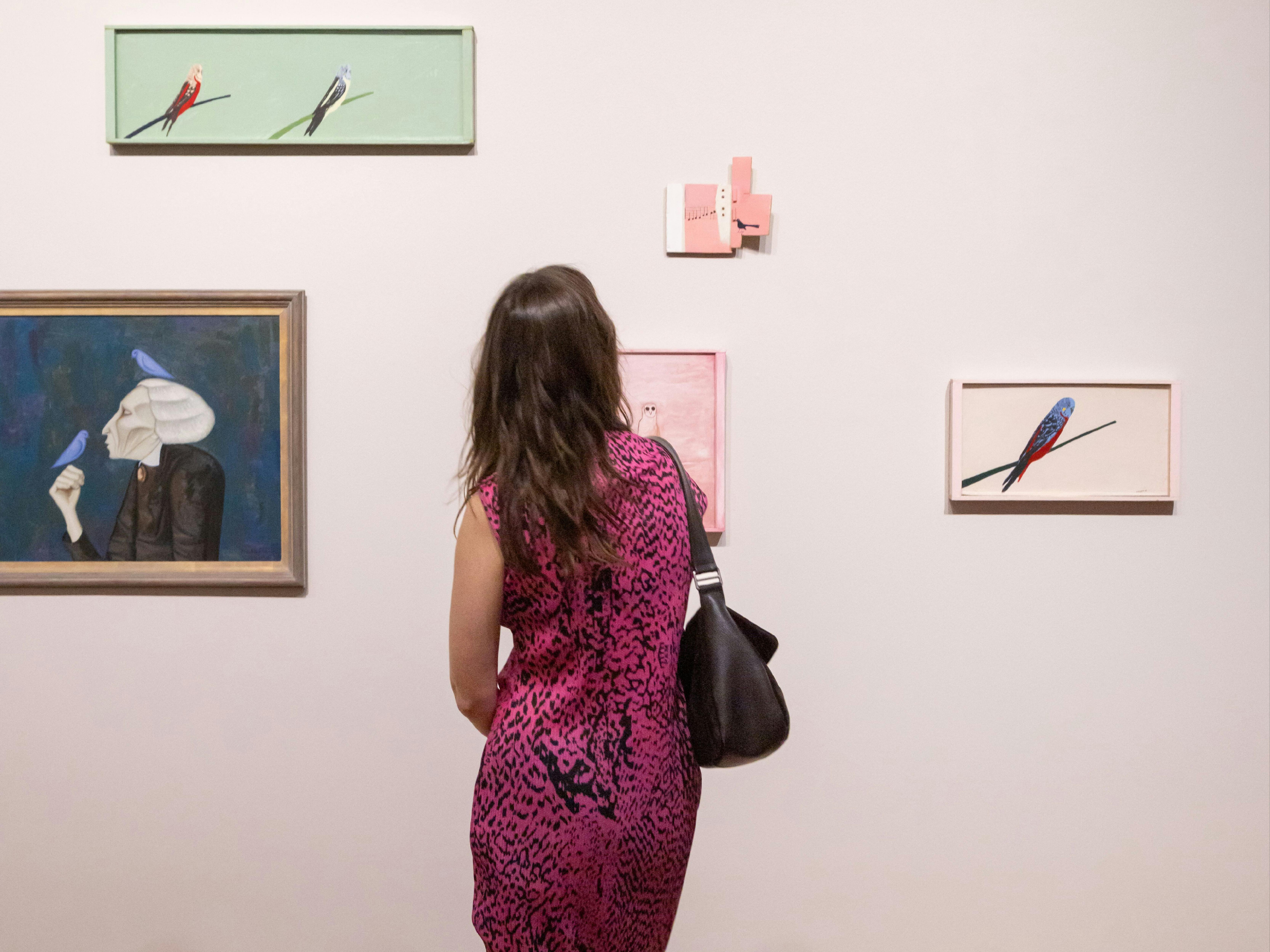 A person wearing a patterned dress views several painted portraits on a light-coloured wall.
