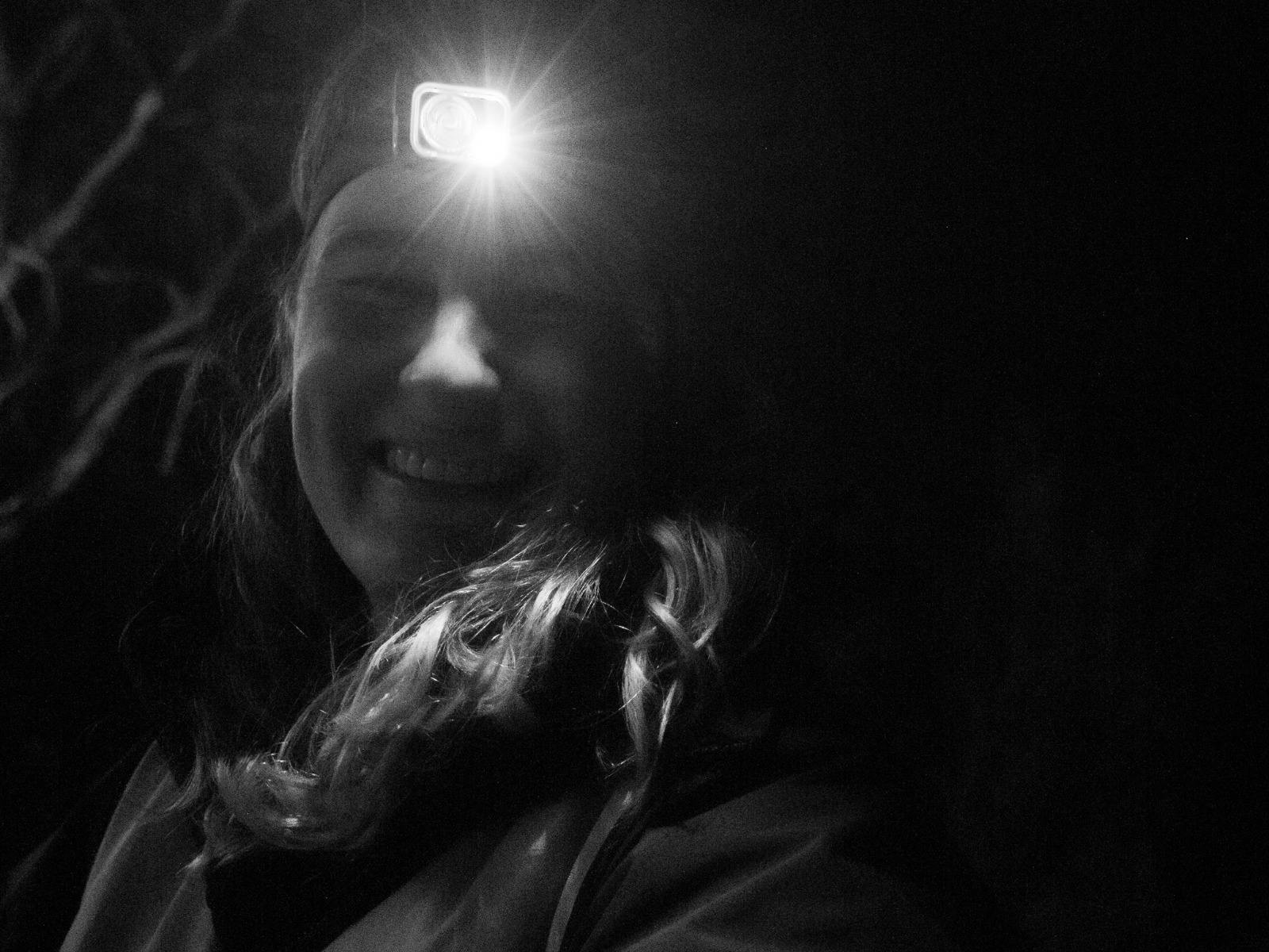 A  black and white photo of a woman wearing a headtorch smiles